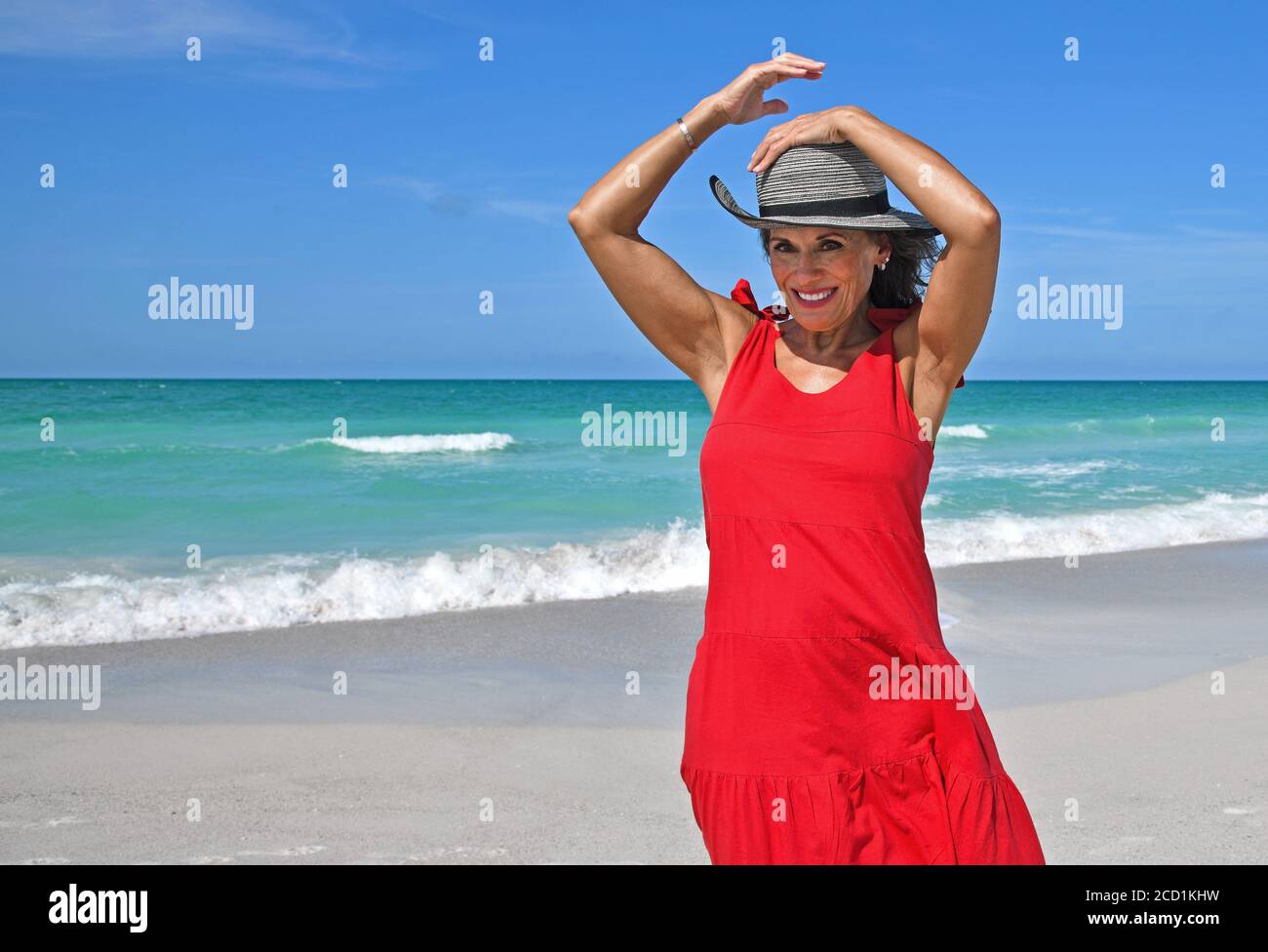 Bella donna matura che indossa un abito estivo rosso sul Spiaggia che tiene il suo cappello nel vento Foto Stock