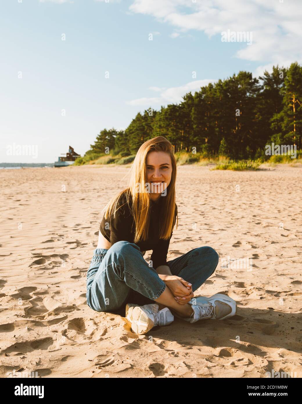 Intera lunghezza bella donna sorridente seduta sulla spiaggia al tramonto in sera. Bella donna con lunghi capelli marroni, guardando la macchina fotografica. Vita lenta, disintossicazione sociale, autoimmersione, essere concetto unico. Foto Stock