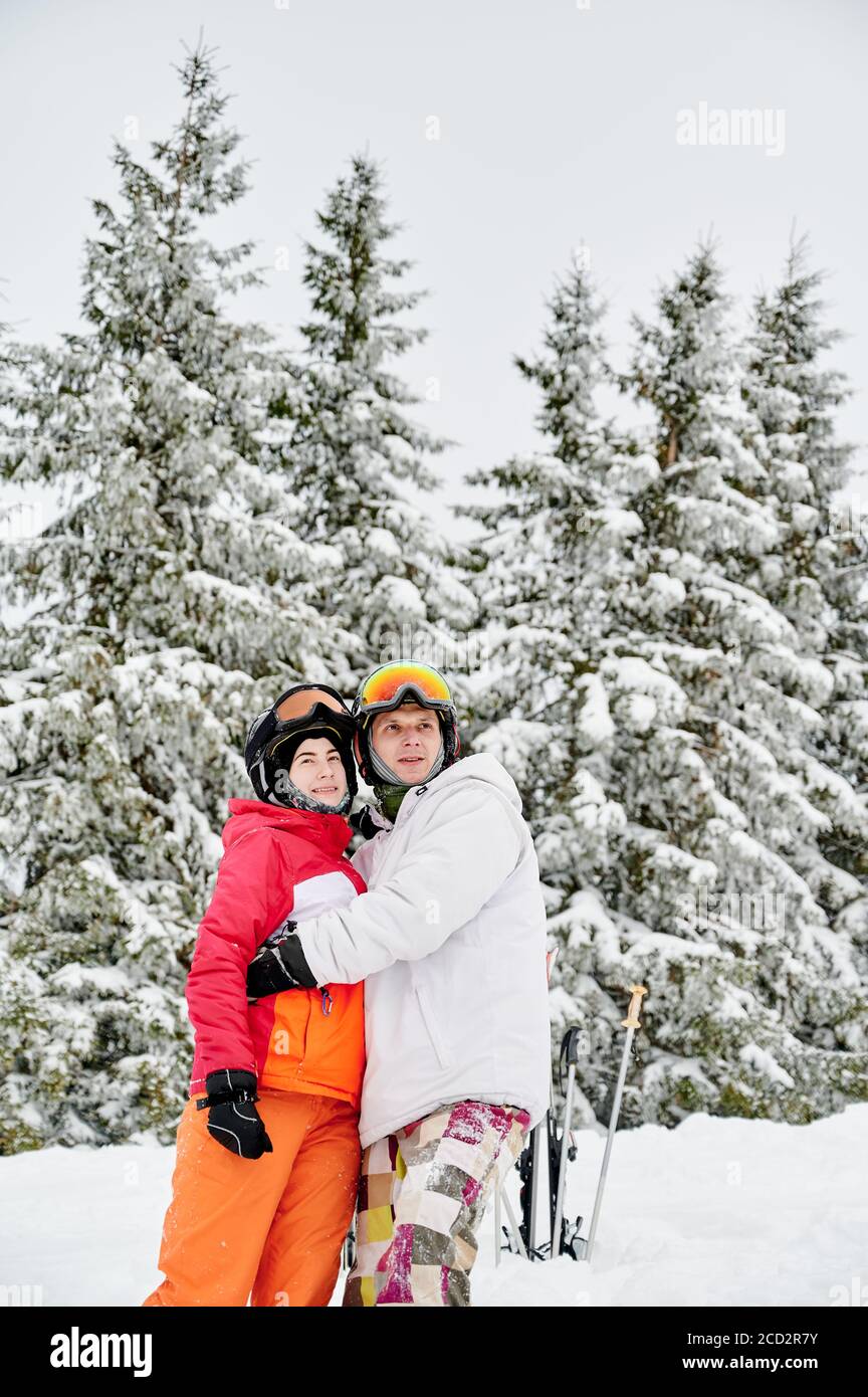 Affascinante uomo e donna in abiti da sci e caschi abbracciano e guardando da parte mentre si trova in piedi nella foresta invernale con alberi di conifere nevosi sullo sfondo. Uomo sciatore abbracciare bella giovane donna e sorridente. Foto Stock