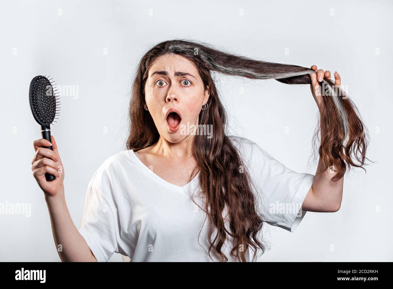 Una donna bruna scioccata tiene un pettine e mostra una ciocca di capelli grigi. Sfondo bianco. Il concetto di perdita dei capelli e capelli grigi. Foto Stock
