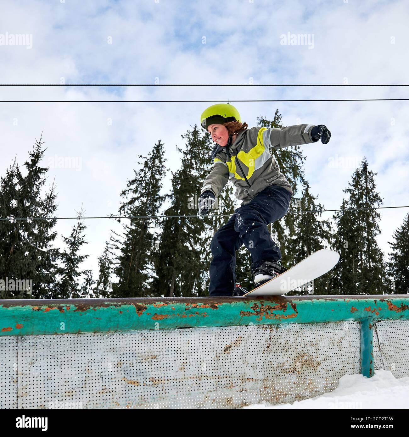 Capretto snowboarder in casco equitazione snowboard sotto il bel cielo nuvoloso con alberi di pino sullo sfondo. Ragazzo che esegue trucchi con snowboard. Concetto di snowboard e attività sportive invernali. Foto Stock