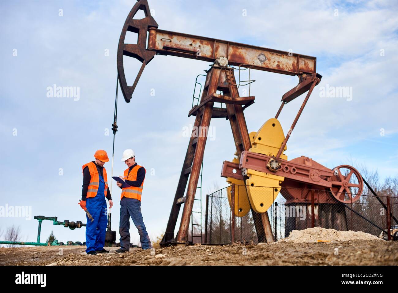 Operatore di estrazione sorridente che scrive sulla clipboard mentre lavora con un collega nel campo dell'olio vicino al martinetto della pompa del petrolio. Lavoratori maschi in caschi, giubbotti da lavoro. Concetto di estrazione dell'olio, industria petrolifera. Foto Stock
