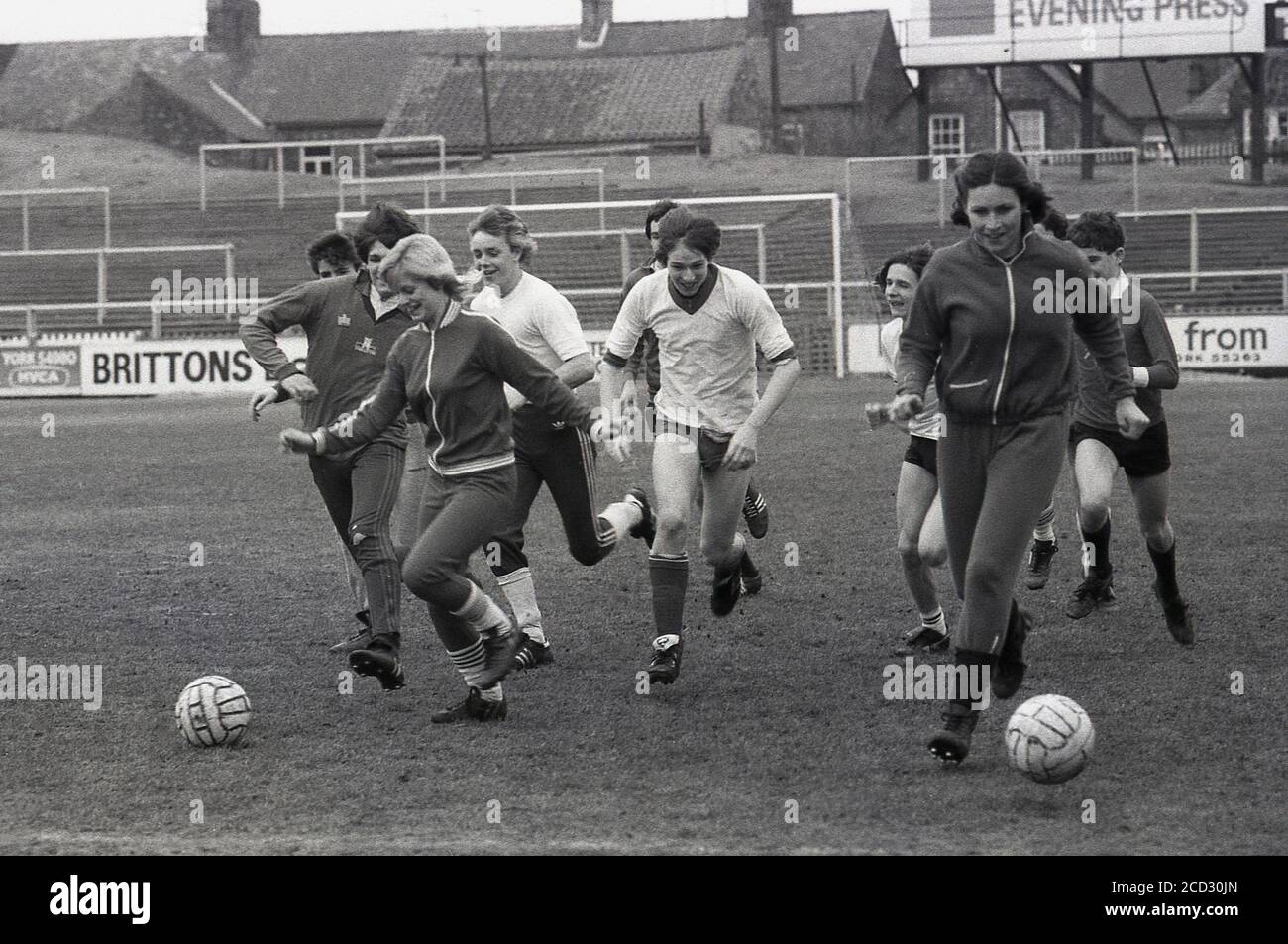 Anni '80, storica, due ragazze in tute da corsa che hanno un calcio sul campo al York City FC sono inseguiti da alcuni ragazzi, tutti i giovani che lavorano a terra avendo un po 'di divertimento prima della partita. Le tradizionali terrazze di calcio erano gli spettatori si levavano in piedi e guardare una partita può essere visto. Foto Stock