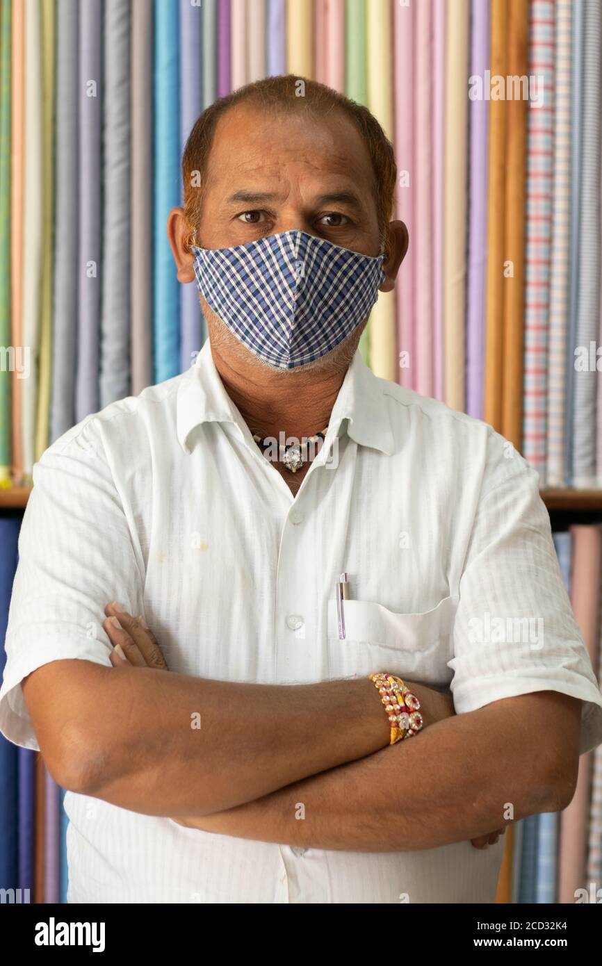 Ritratto del negoziante che guarda la fotocamera in un negozio di stoffa che indossa maschera medica protettiva per prevenire il coronavirus o covid-19 e in piedi con bracci Foto Stock