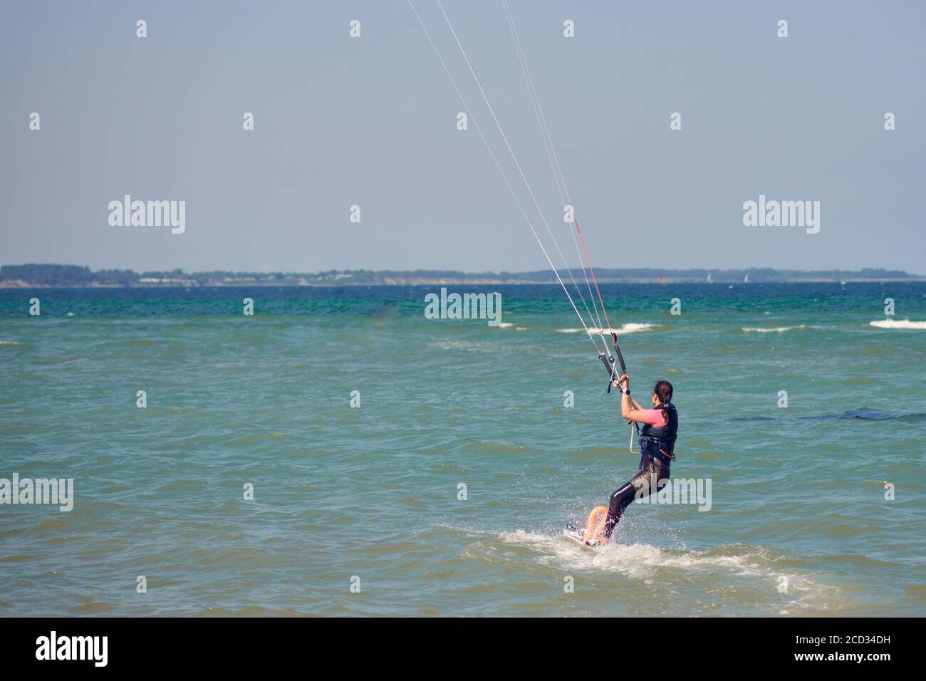 Bruna donna kitesurf o kite boarding che si allontana dal spiaggia di sabbia che fa per acque più profonde in un'estate soleggiata giorno in una vista posteriore del venuto Foto Stock