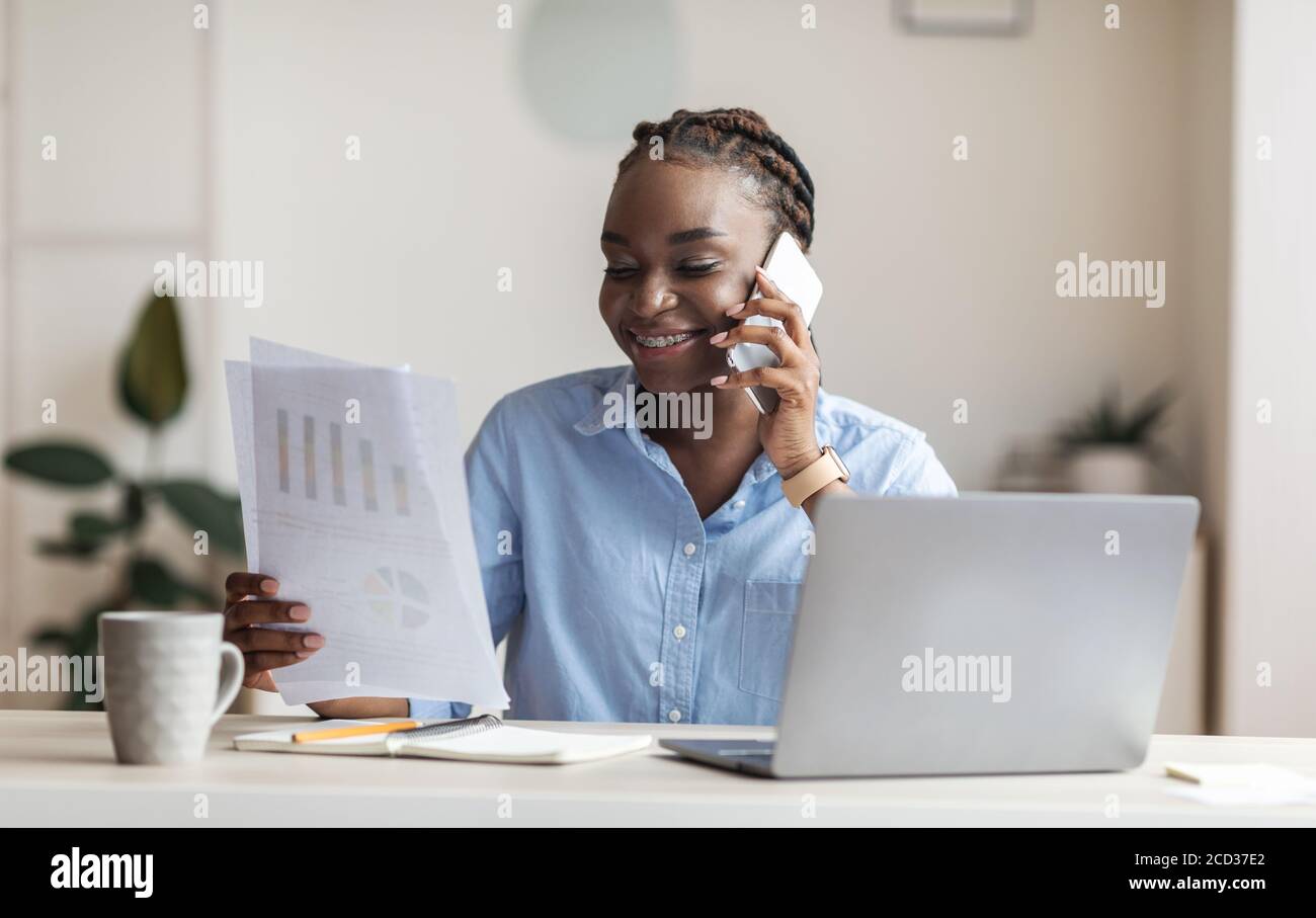 Donna d'affari nera riuscita che controlla i rapporti finanziari e che parla sul cellulare in ufficio Foto Stock
