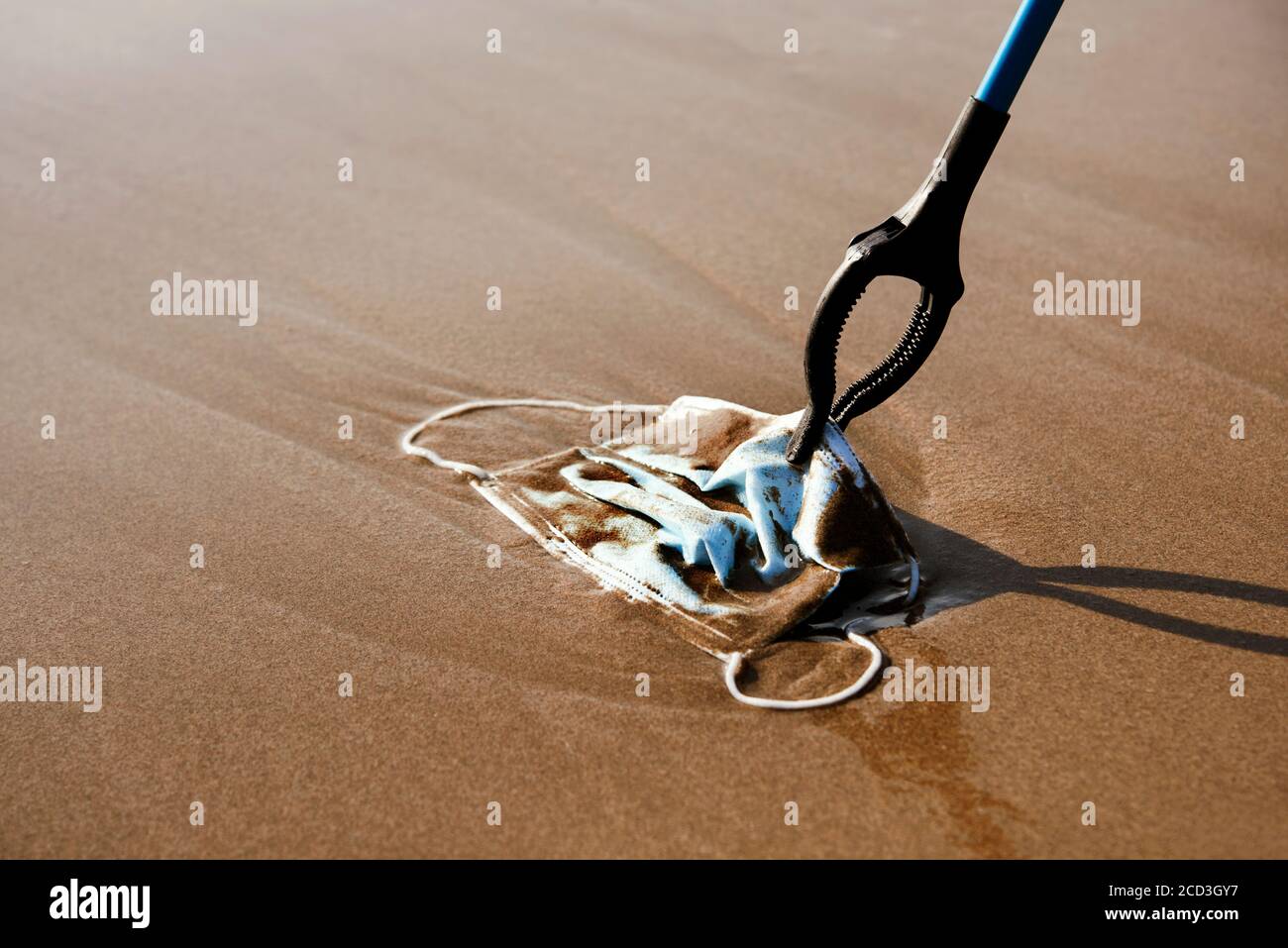 closeup di qualcuno che utilizza un estensione reach per raccogliere un maschera chirurgica blu utilizzata gettata sulla sabbia bagnata di il mare o riportato dall'oceano a th Foto Stock