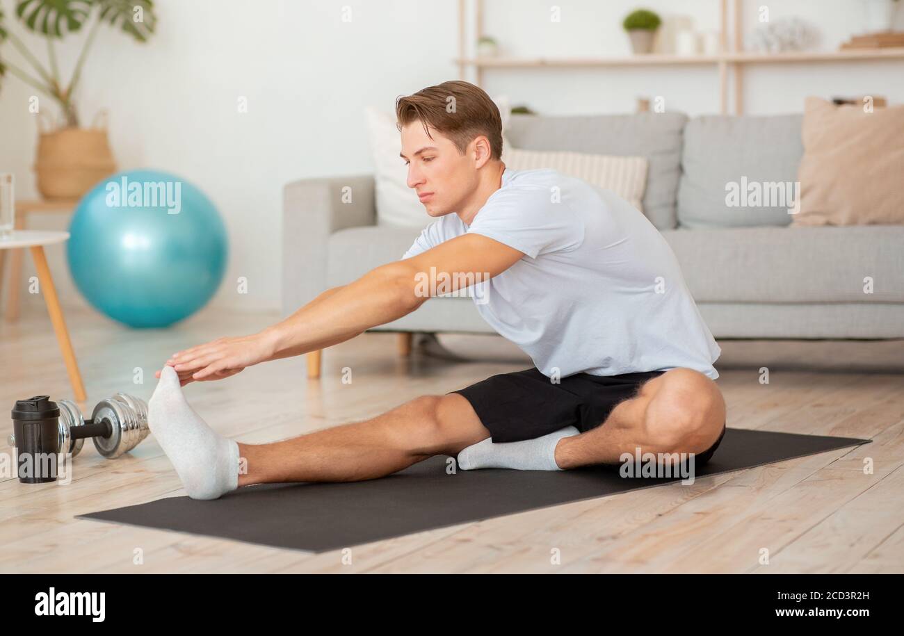 Tempo libero e l'esercizio a casa durante la quarantena. L'uomo muscoloso che fa stretching per le gambe sul tappetino Foto Stock
