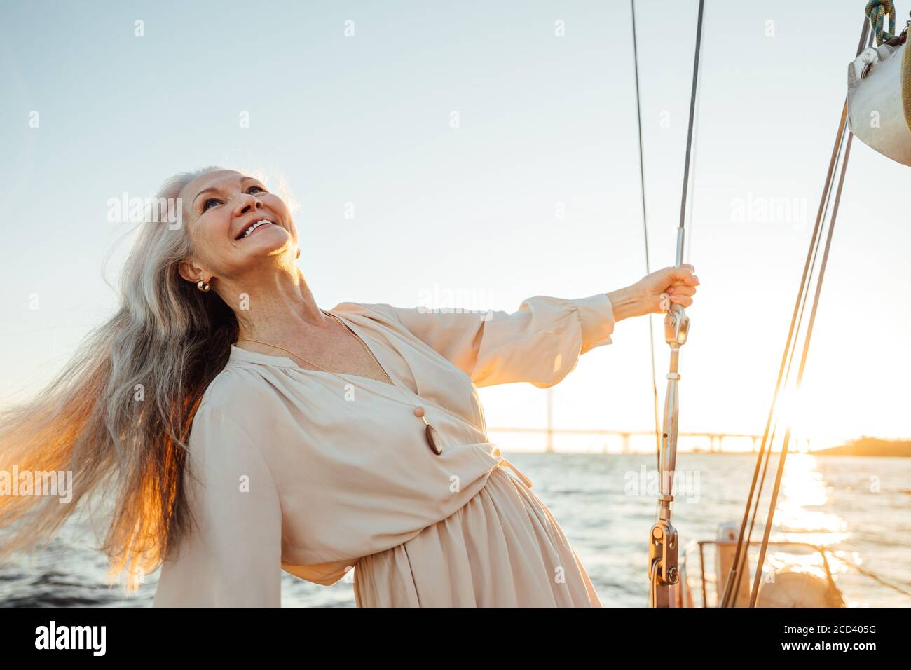 Bella donna matura con capelli lunghi godendo il tramonto. Donna sorridente che tiene una corda e guarda in su mentre si alza su una barca a vela. Foto Stock