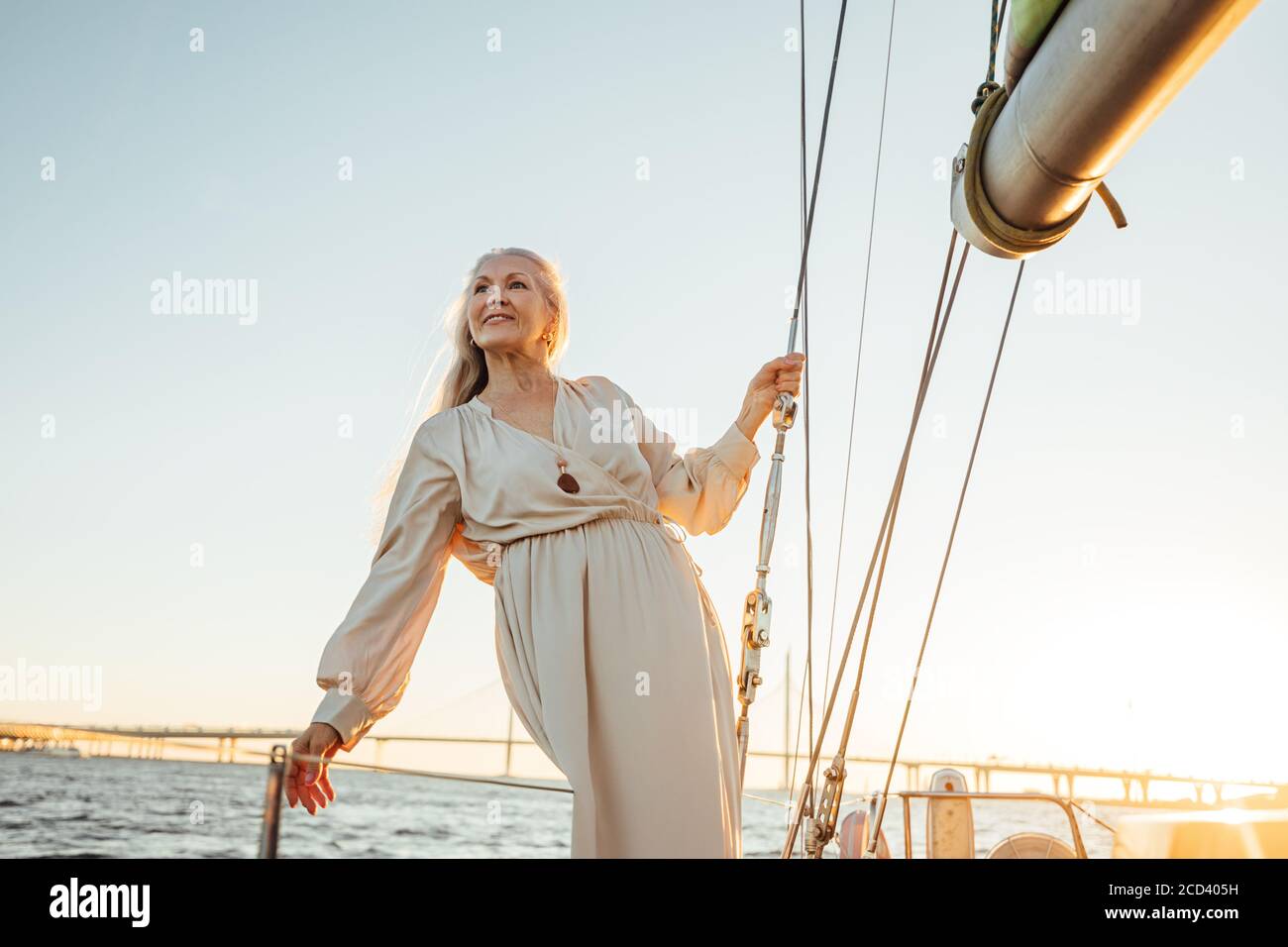 Ritratto di donna matura in abito in piedi sullo yacht al tramonto. Bella donna anziana che tiene una corda e che guarda via. Foto Stock