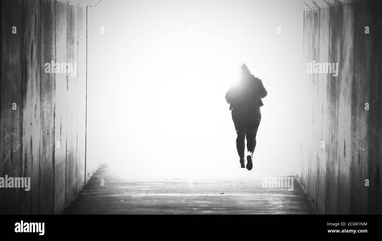 Una figura femminile isolata e solitaria svanisce dirigendosi o camminando verso una luce luminosa alla fine del tunnel. Dolore, morte, suicidio morente, concetto di fede. Foto Stock