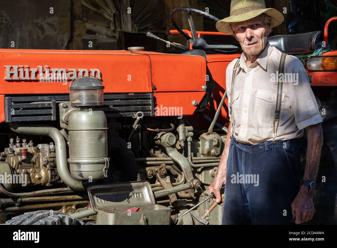 Vecchio agricoltore che lavora su un trattore rosso sotto il tetto della casa contadina sotto il sole. Vintage, vista laterale. Foto Stock