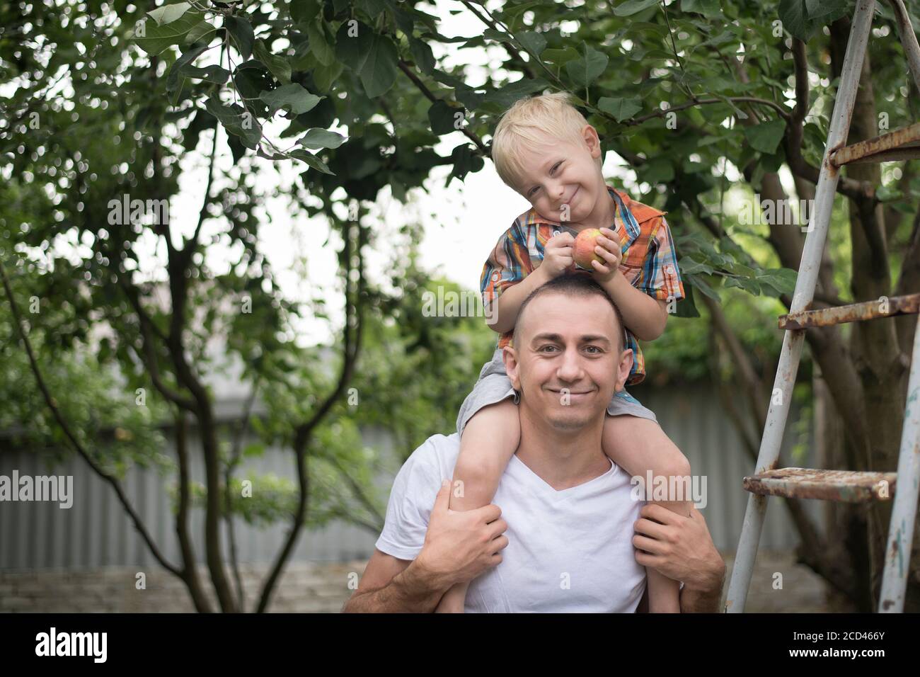 Ragazzo biondo con una mela nelle mani sulle spalle del padre. Giardino sullo sfondo Foto Stock