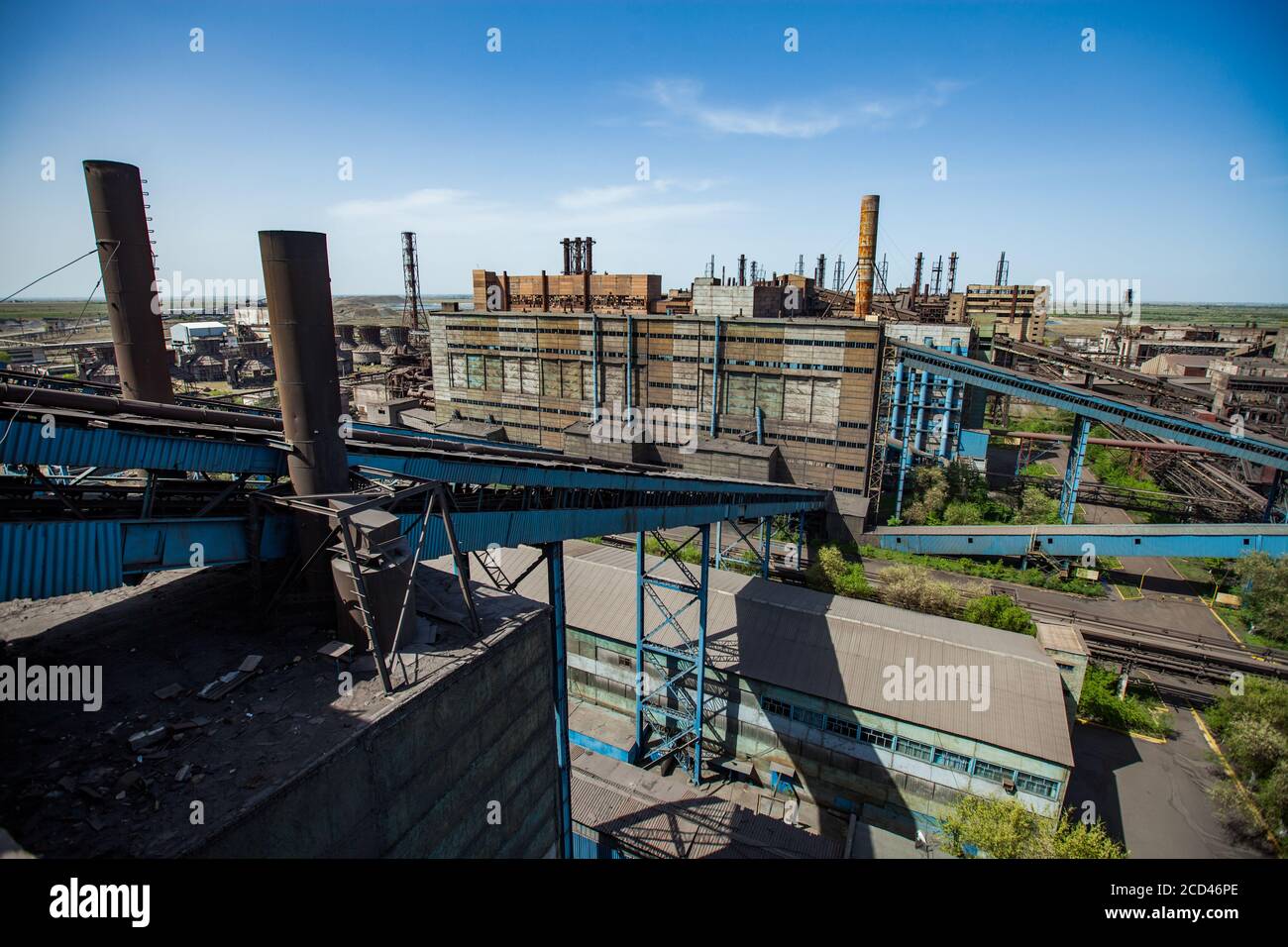 Impianto di metallurgia sovietica. Edifici di fabbrica abbandonati e trasportatori blu e camini arrugginiti sul cielo blu. Vista dall'alto (aerea). Taraz Foto Stock