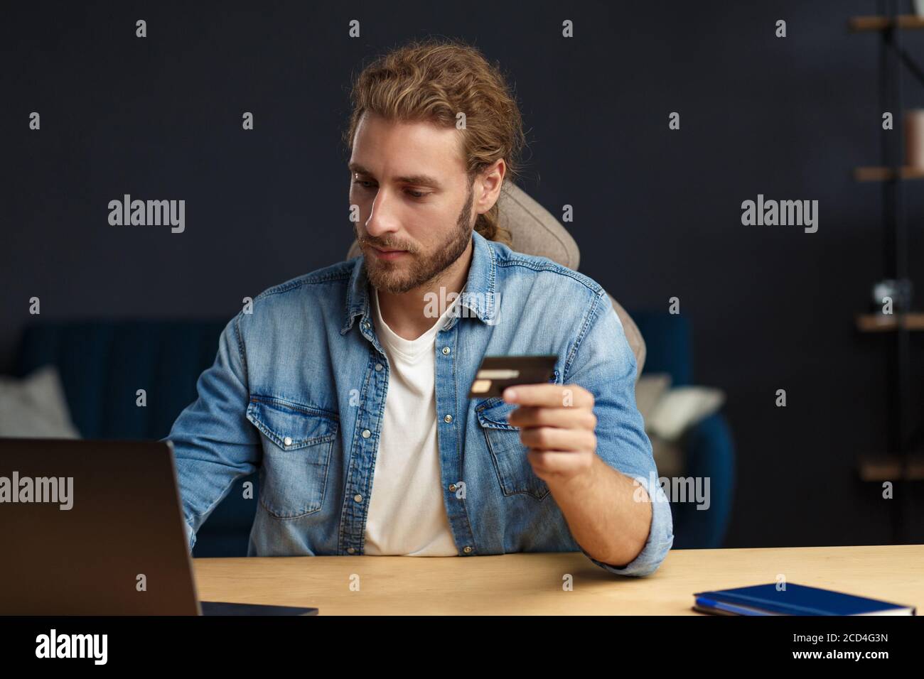 Giovane bell'uomo sorridente riccamente con capelli lunghi in ufficio di casa tenendo la carta di credito e utilizzando il computer portatile. Acquisti online, pagamenti online. Prenotazione biglietti Foto Stock