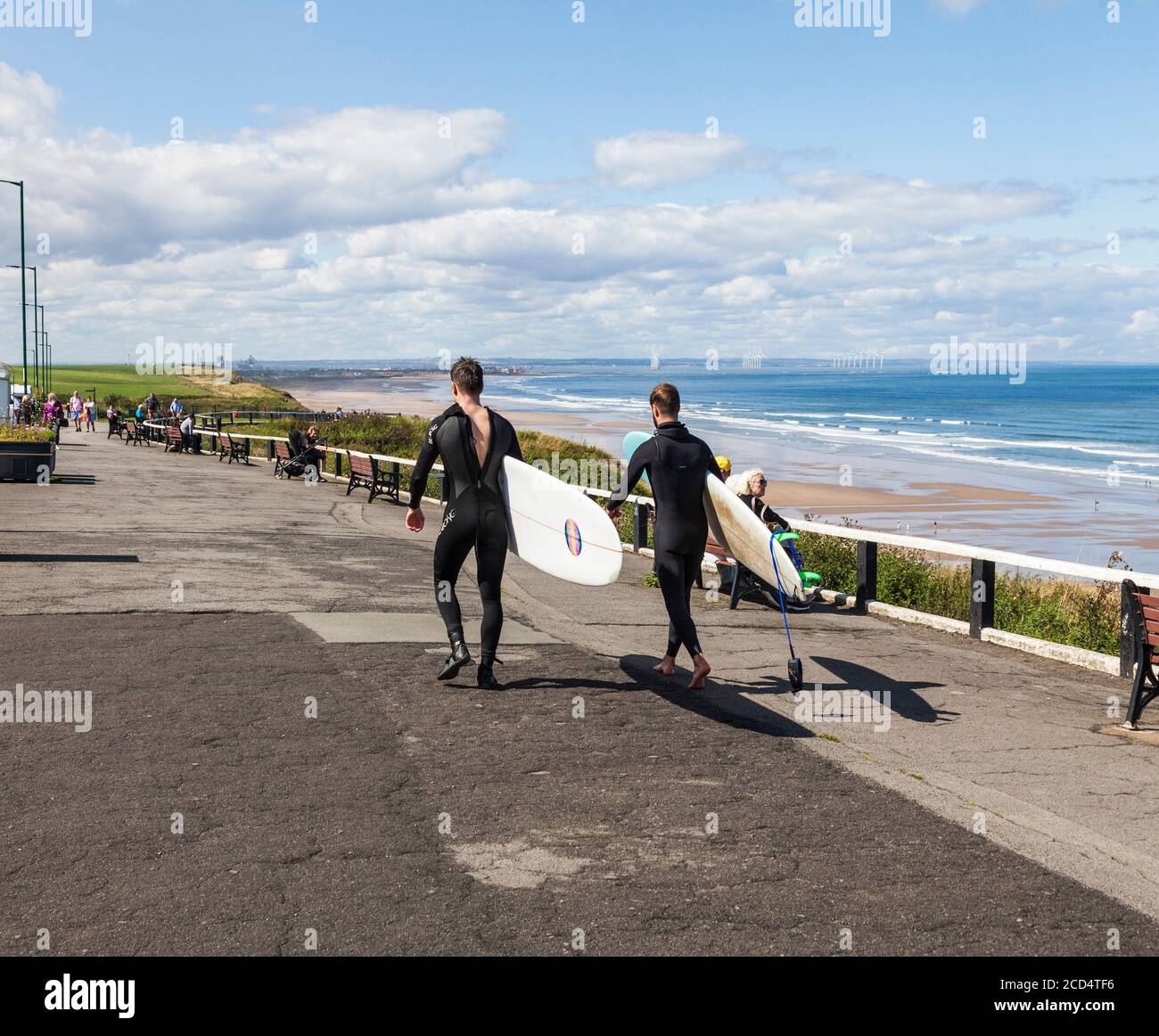 Due giovani uomini in tute umide che trasportano le loro tavole da surf Lungo la passeggiata a Saltburn by the Sea, England, UK Foto Stock