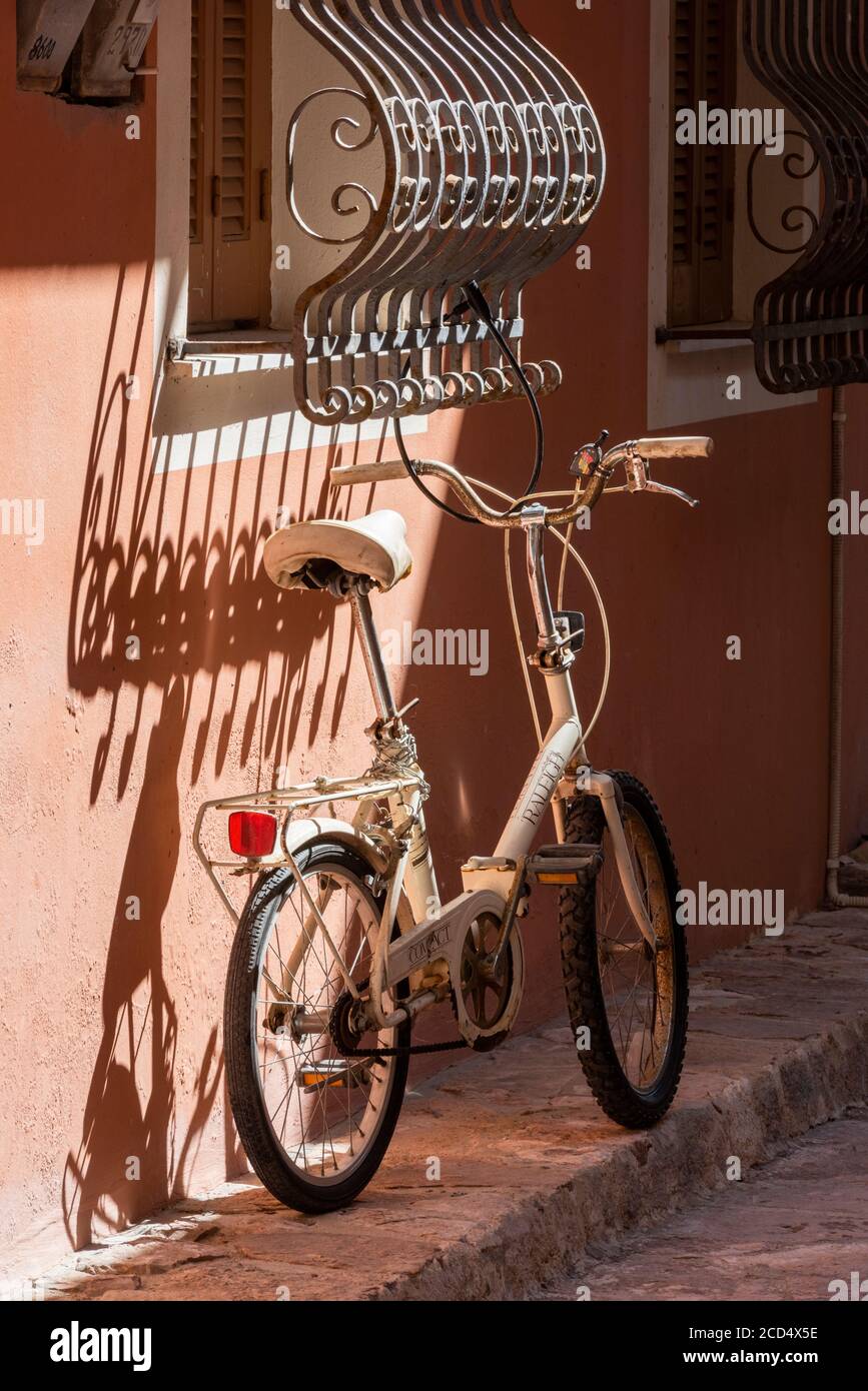 una bicicletta pieghevole appoggiata al muro di una serata estiva con ombre che cadono su un muro e persiane in una città greca a zante. Foto Stock