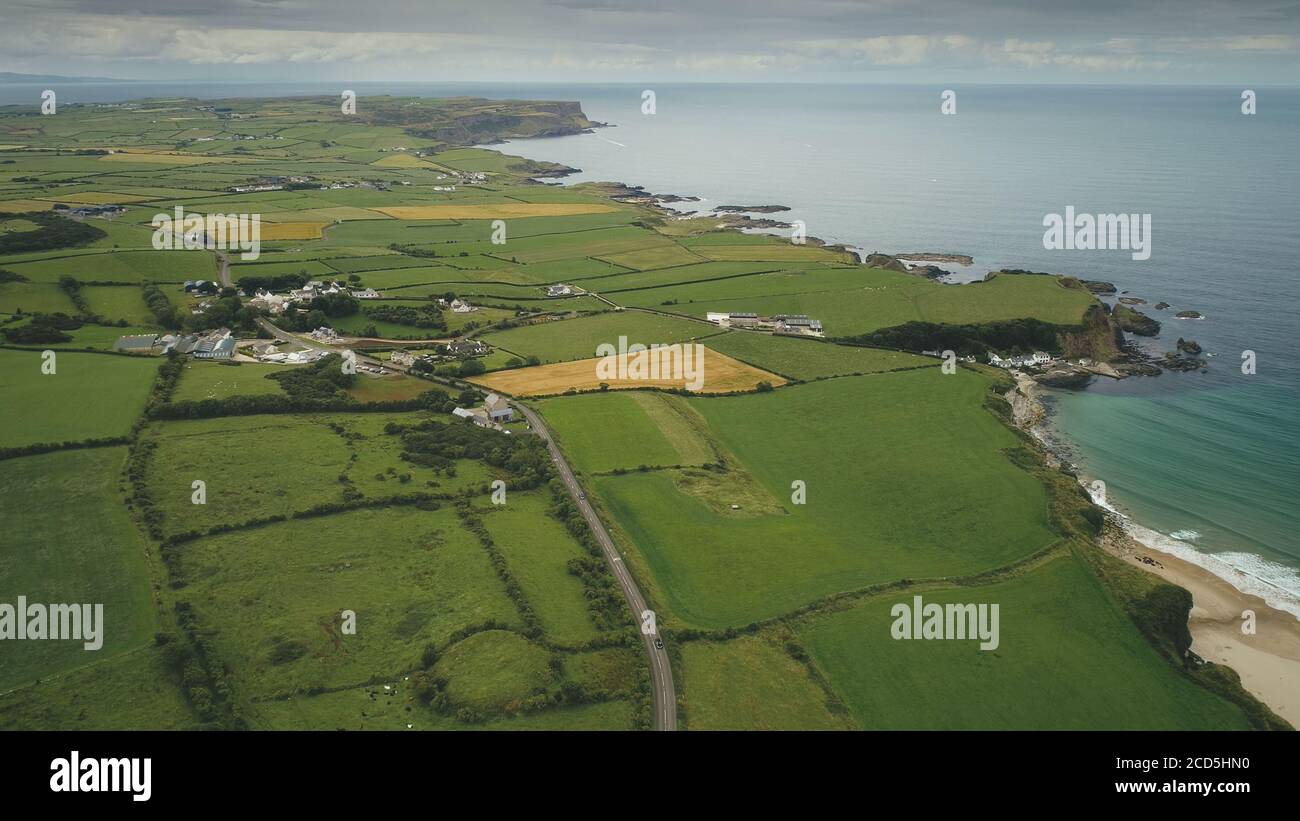Irlanda aereo verde campi paesaggio shot: Strada lungo prati. Ampie piante e fattorie sulla costa rocciosa dell'oceano Atlantico. Scenario epico dell'Isola d'Irlanda del Nord. Scatto panoramico Foto Stock