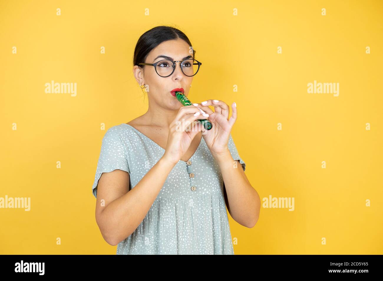 Giovane bella donna che indossa occhiali in piedi su isolato sfondo giallo riproduzione flauto Foto Stock