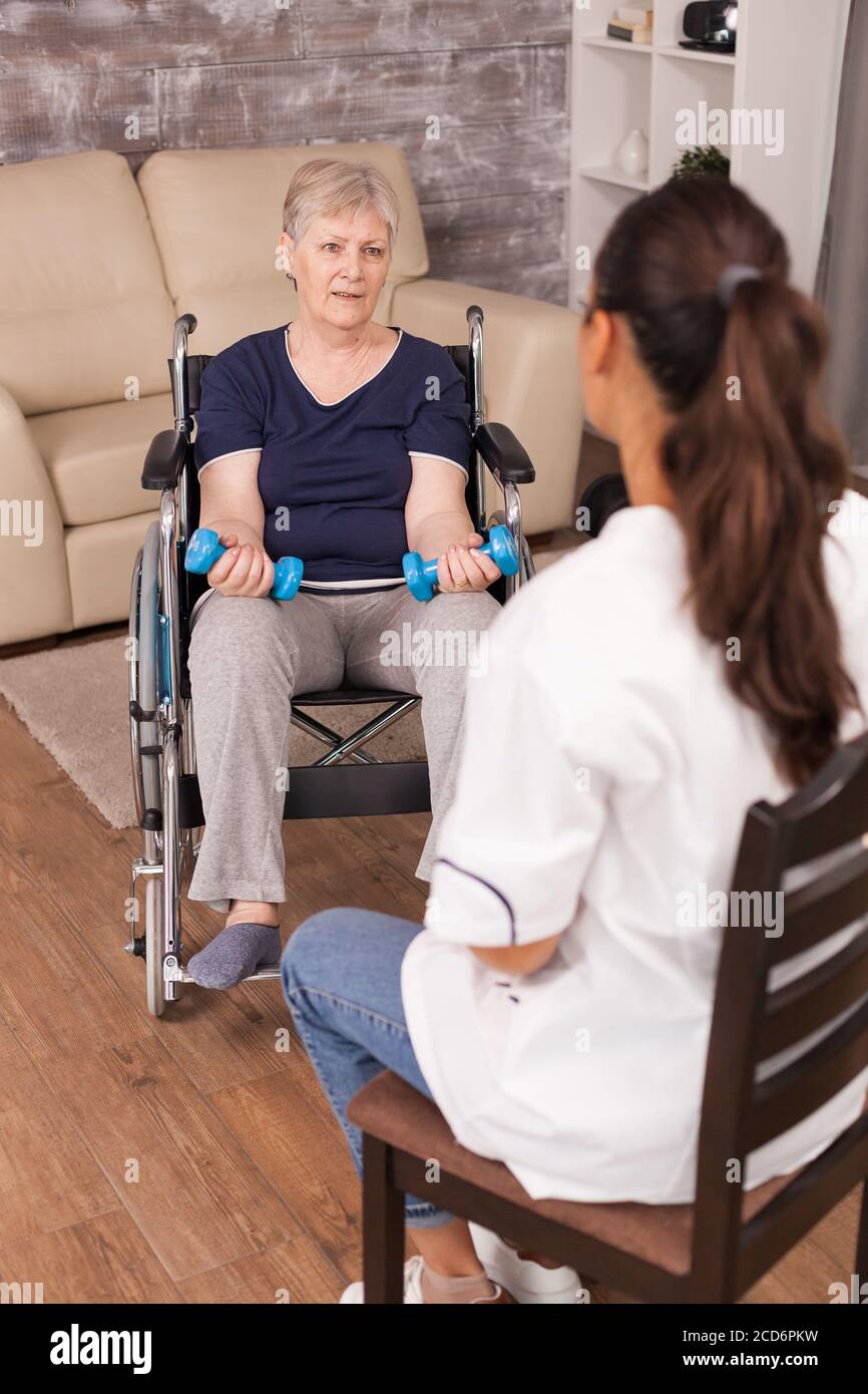 Donna in sedia a rotelle avere una conversazione con nurseNurse spiegare esercitazioni di dumbbell anziano disabile che recupera l'infermiera di aiuto professionale, trattamento di pensione di cura e riabilitazione. Foto Stock