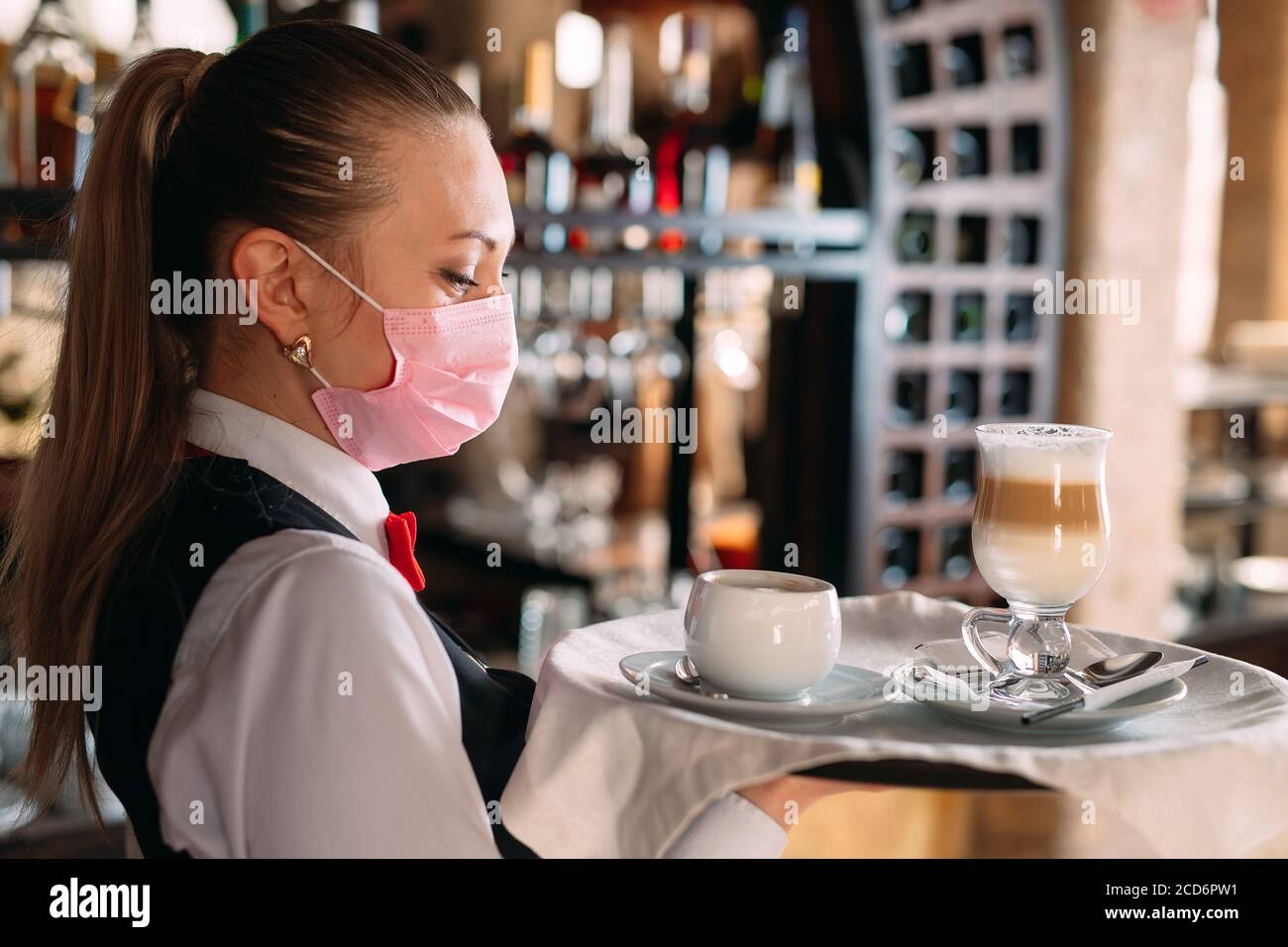 Una cameriera femminile di aspetto europeo in una maschera medica serve caffè latte. Foto Stock