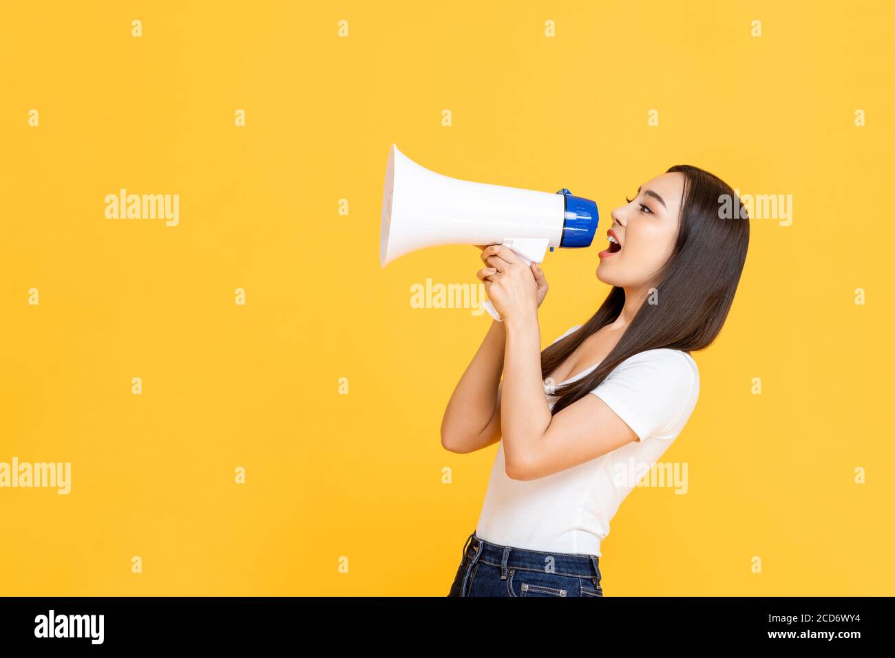Vista laterale ritratto di giovane bella donna asiatica che tiene il megafono mentre gridando annuncio in studio isolato sfondo giallo Foto Stock
