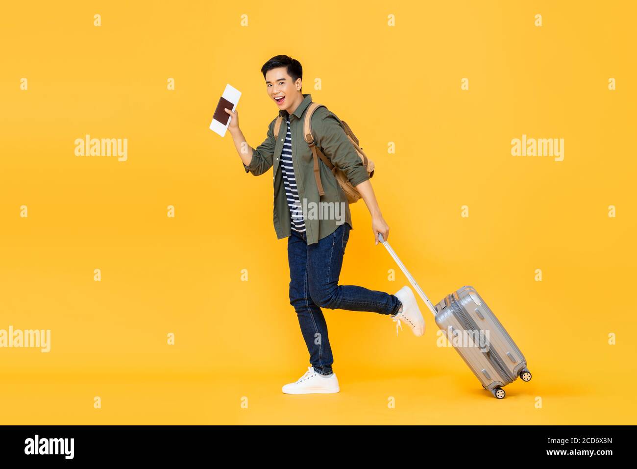 Ritratto a lunghezza intera di allegro giovane sorridente turista asiatico uomo camminando tenendo in mano i bagagli e mostrando il passaporto con il biglietto in entrata studio isolato yel Foto Stock