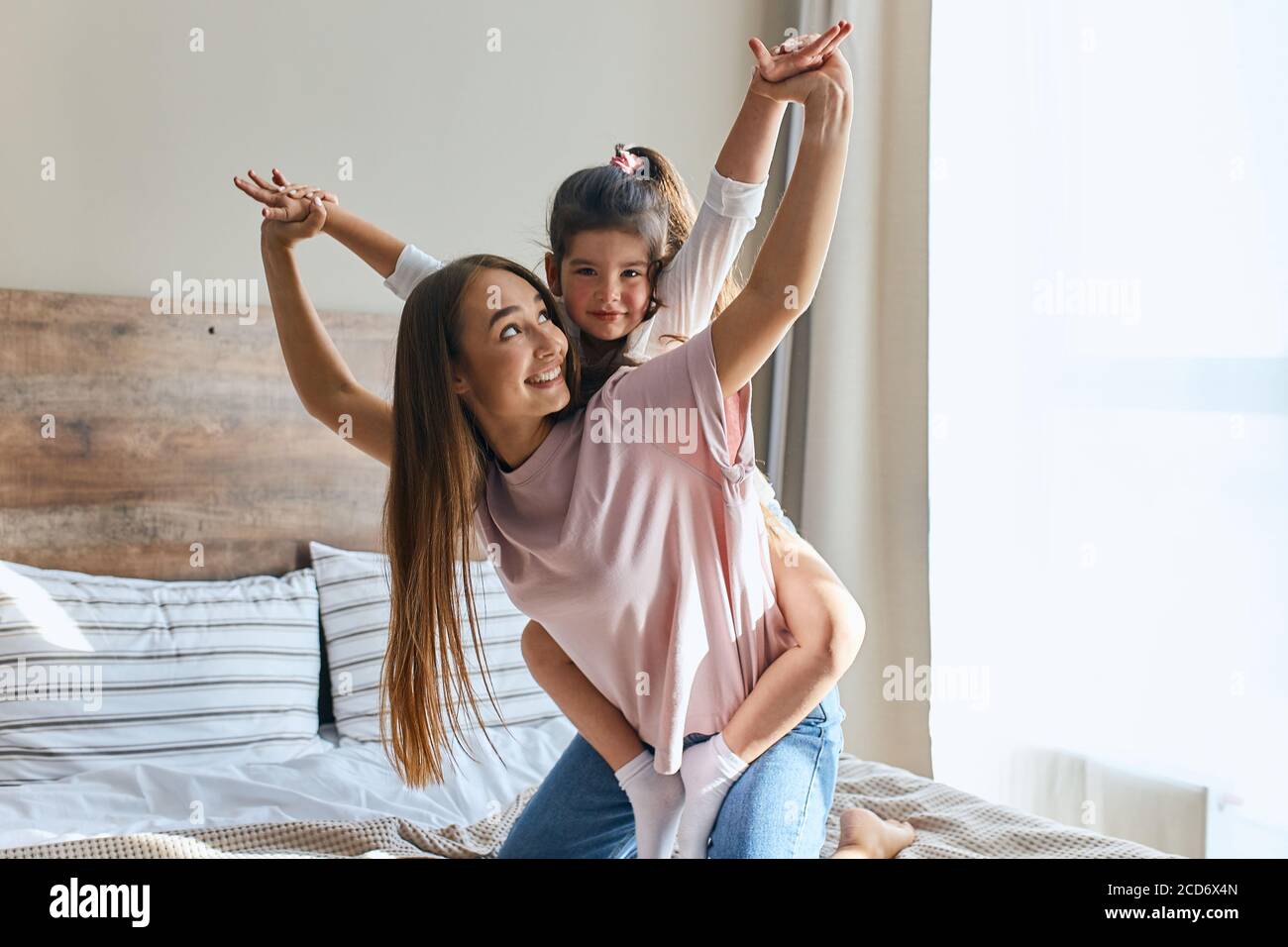 Affascinante allegra ragazza di quattro anni seduta sulla parte posteriore di mamma giocosa, guardando la macchina fotografica, spargendo le teste lateralmente, felice di essere con mamma, caldo e c Foto Stock