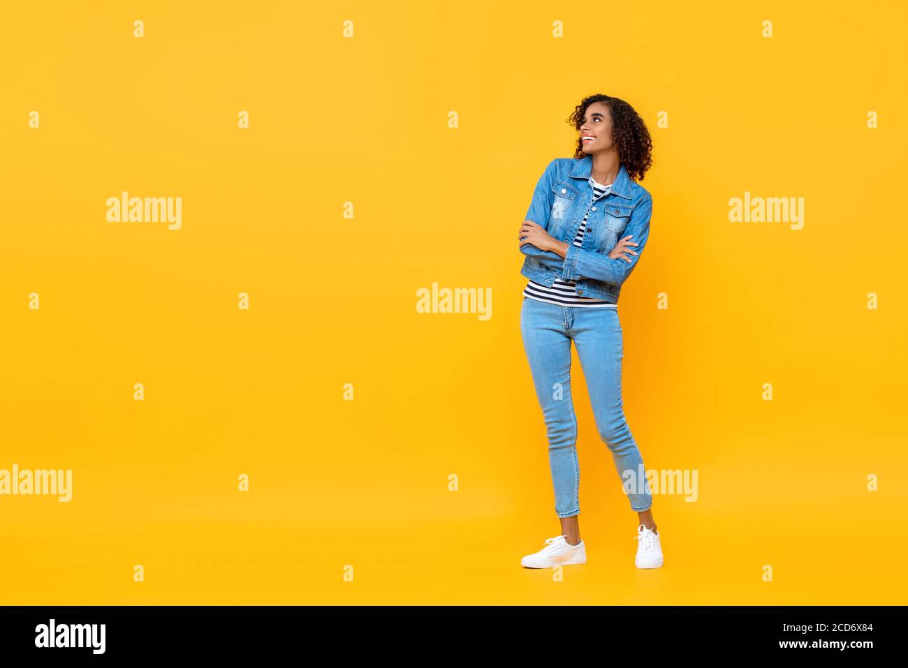 Ritratto a lunghezza intera di giovane donna afroamericana sorridente che guarda a lato con le braccia incrociate su sfondo studio giallo Foto Stock