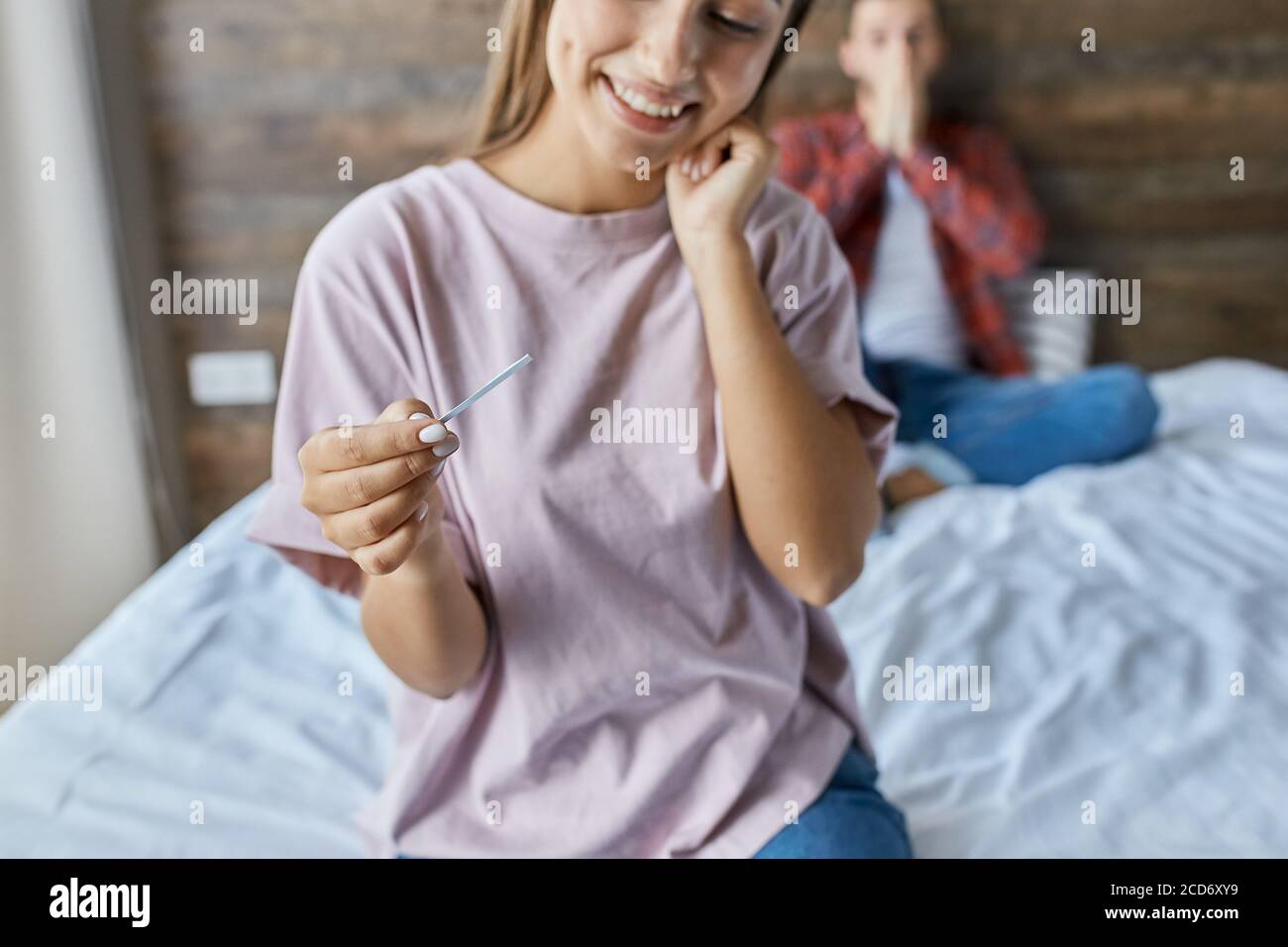 Allegra donna felice fissa al test di gravidanza con emozioni positive, guardando via con ampio sorriso e volto felice, sfondo offuscato di uomo scioccato, Foto Stock