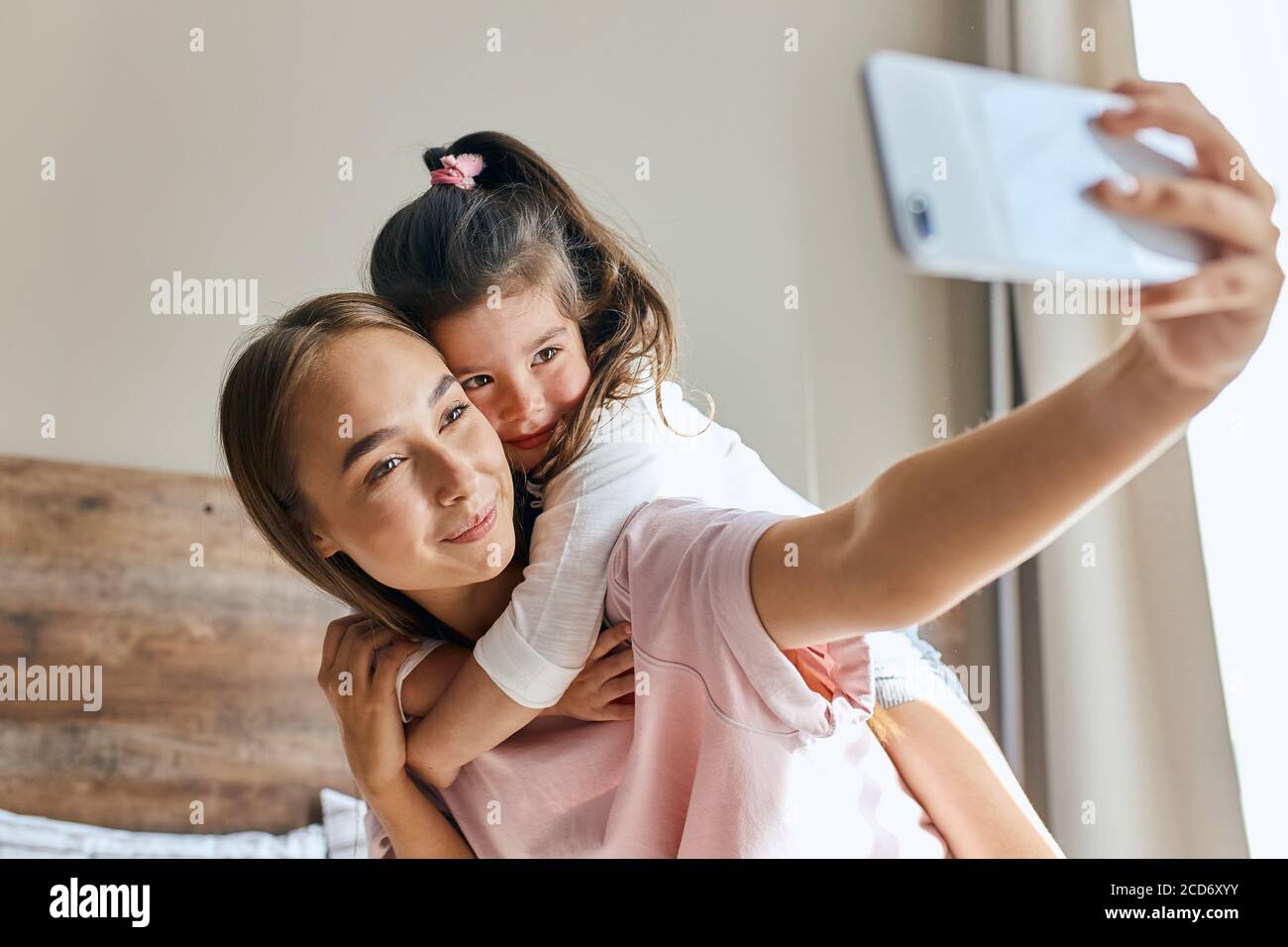 Affascinante giovane donna prende selfie utilizzando il telefono cellulare, carino allegro bambino hugs mamma, esprimendo la felicità, guardando da parte, si concentra in dispositivo, primo piano, pos Foto Stock