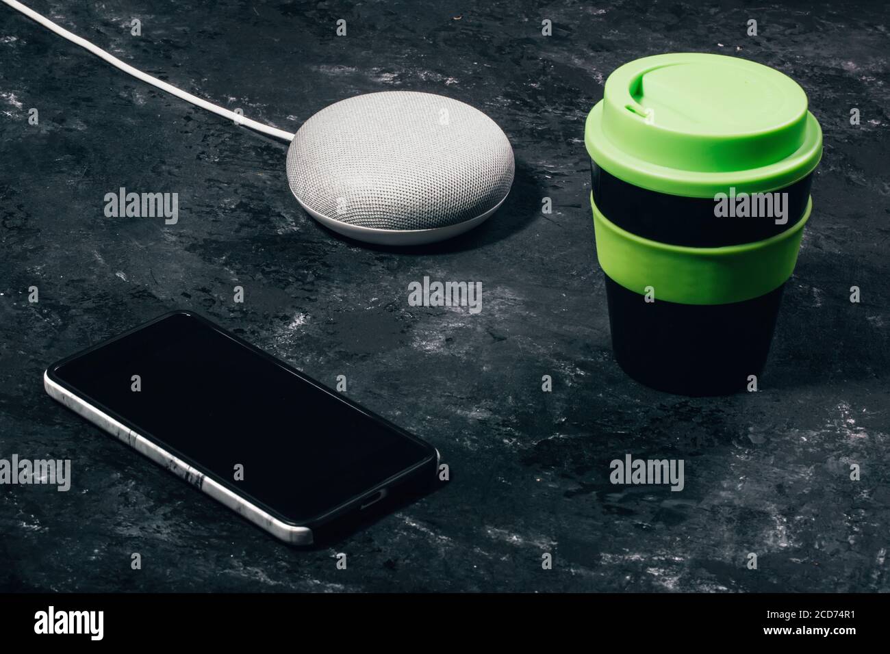 Concetto di assistente digitale. Altoparlante intelligente con luci a LED attivate su un tavolo nero in cemento con smartphone e una tazza da caffè riutilizzabile. Foto Stock