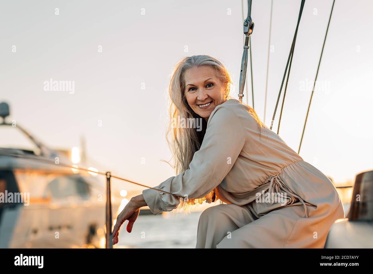 Donna giovane allegra seduta sul ponte dello yacht e guardando la macchina fotografica. Buona donna anziana che si gode fine settimana di viaggio in acqua. Foto Stock