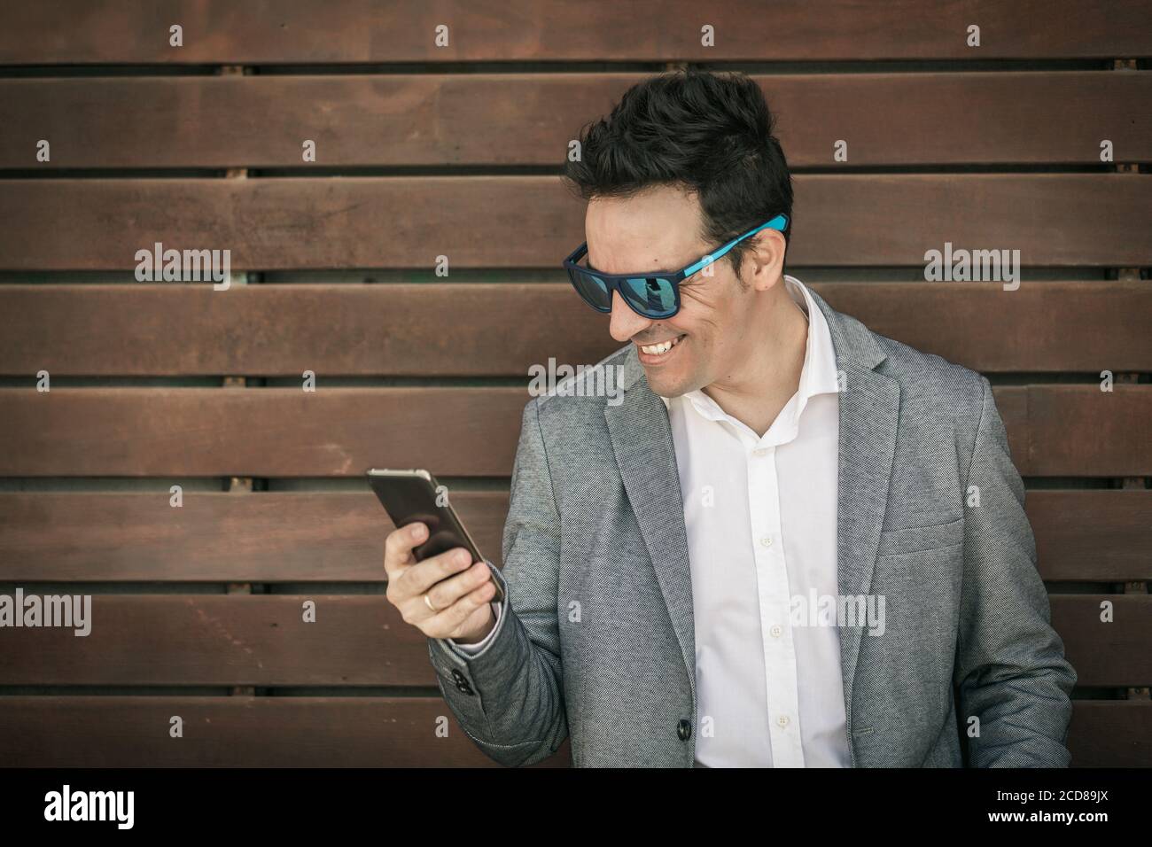 Felice uomo di stile in abito elegante e occhiali da sole alla moda in piedi contro il muro di legno e la lettura di notizie su internet via smartphone Foto Stock