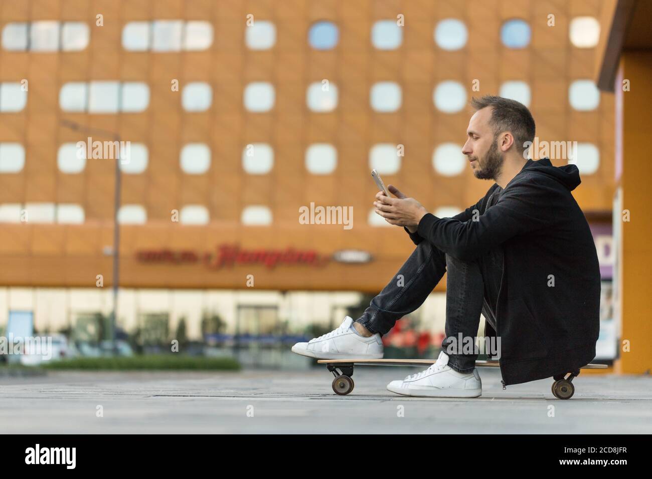 Giovane uomo hipster in abiti neri casual con smartphone, riposo, seduta su tavola lunga o skateboard all'aperto, vista laterale Foto Stock