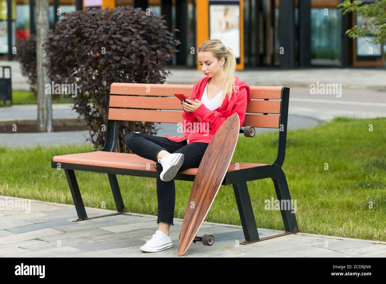 Giovane donna seduta con skateboard o longboard su panca di legno, con smartphone, riposo. Foto Stock