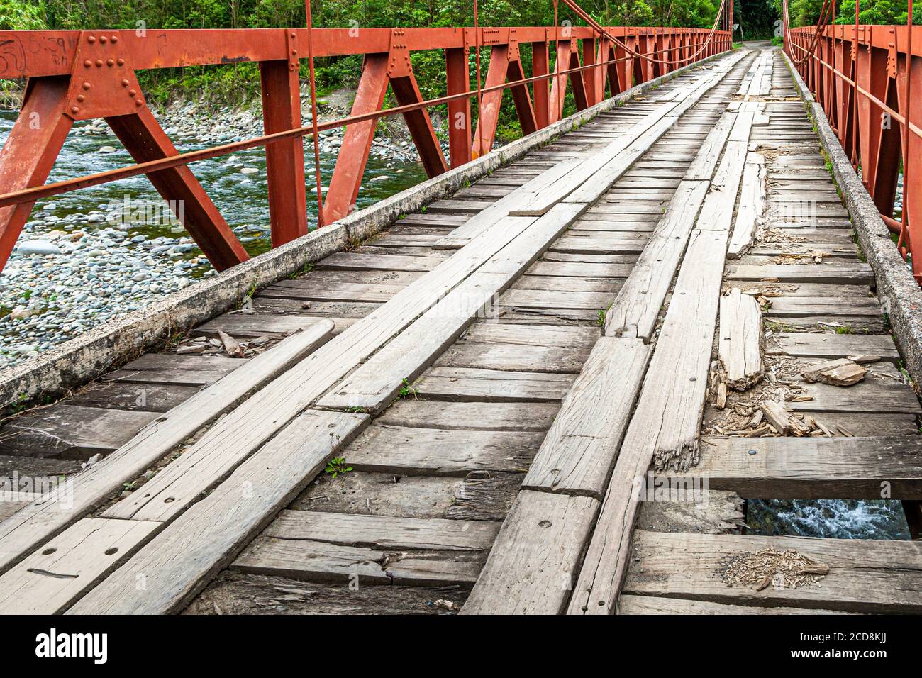La struttura in acciaio dipinta di rosso di un ponte a traliccio si estende su un fiume roccioso nella valle di Orosi, la vista si dirige lateralmente sul letto stretto del fiume con rive di ghiaia. Costa Rica. Foto Stock
