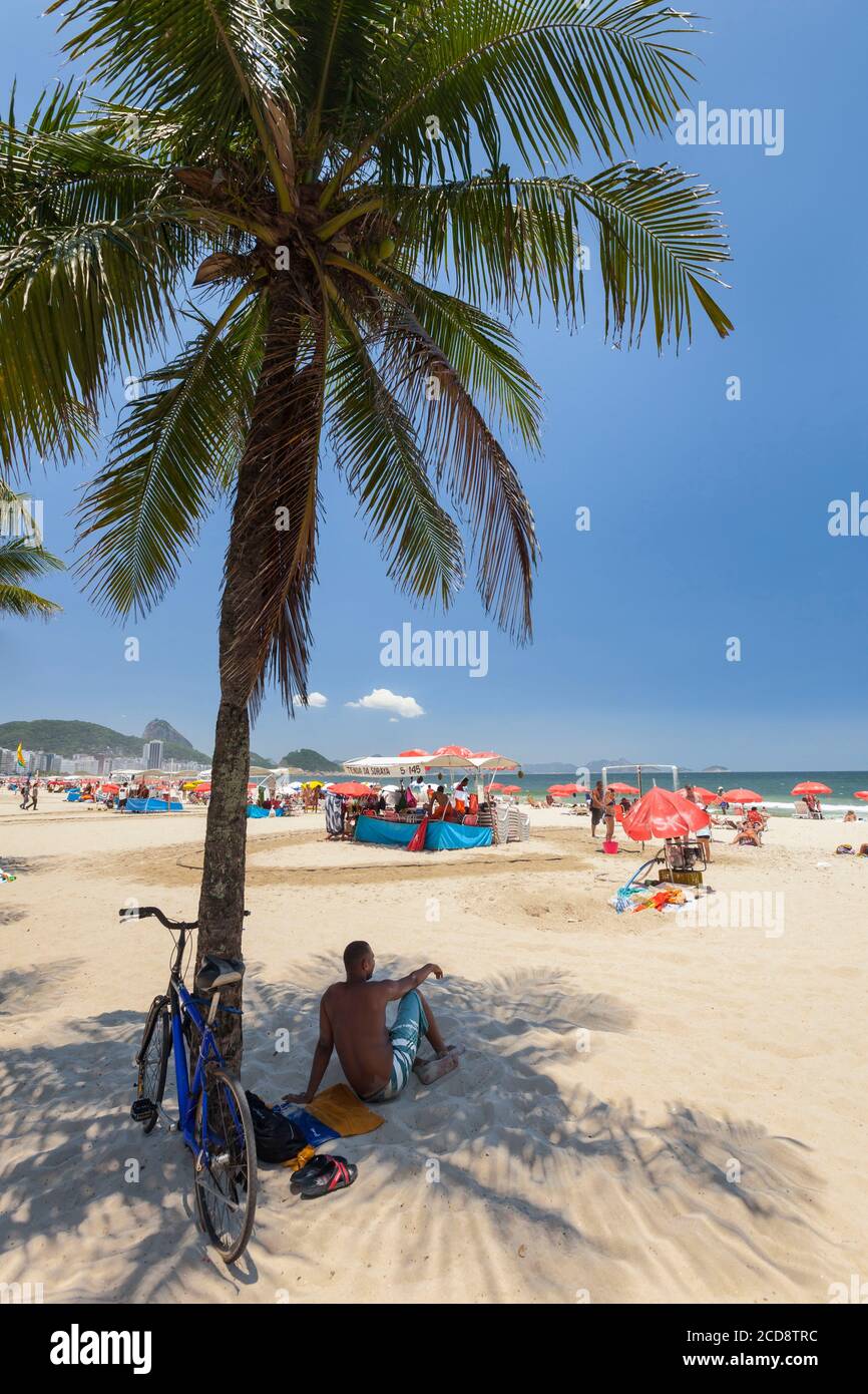 Brasile, Rio de Janeiro stato, Rio de Janeiro città, Copacabana spiaggia, Carioca paesaggi tra la montagna e il mare classificato patrimonio mondiale dell'UNESCO, uomo seduto sulla sabbia, in un'ombra di cocco e guardando la spiaggia Foto Stock