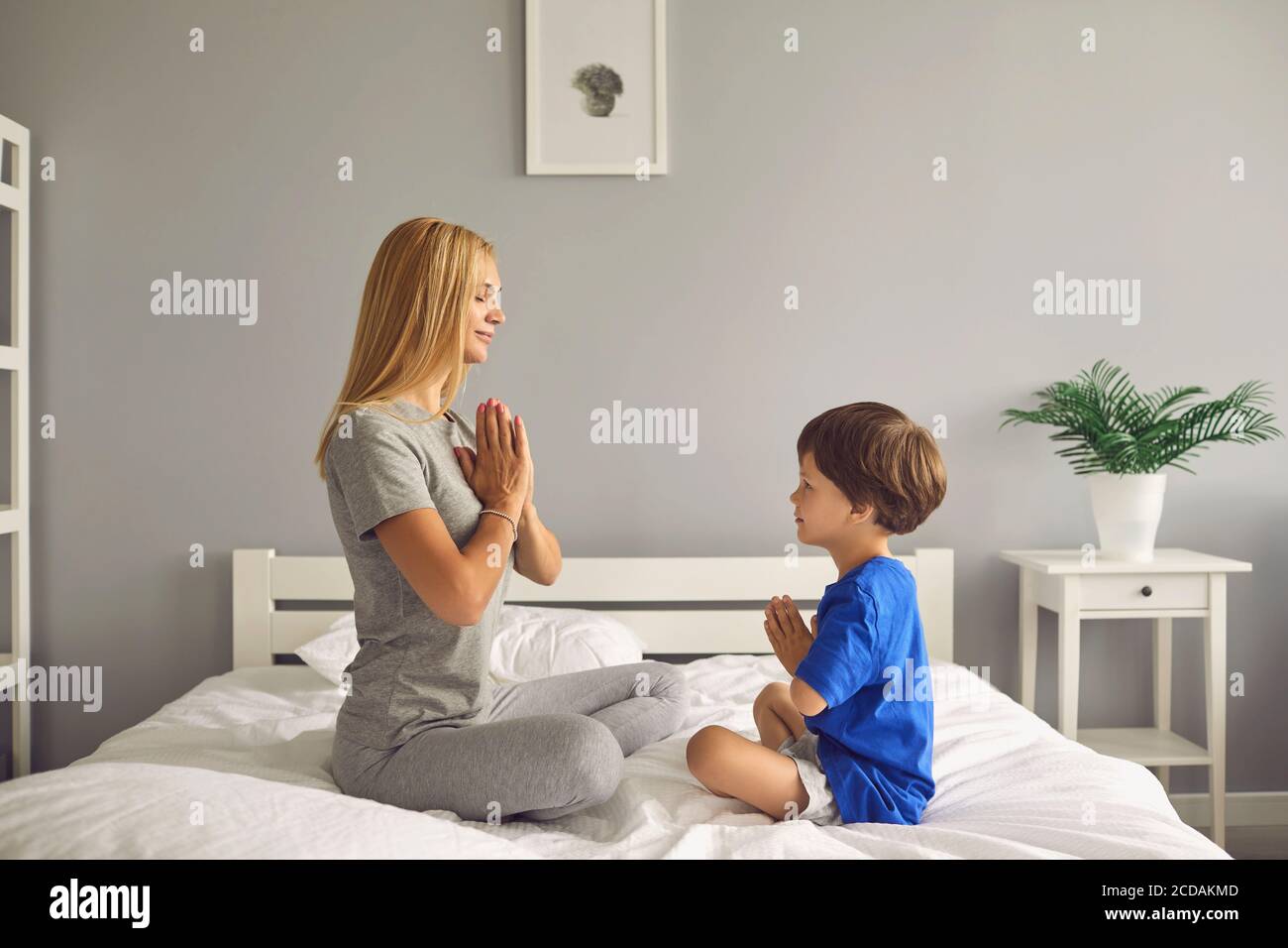 Donna e suo figlio sorridono mentre si siedono in posizione di loto sul letto e meditano a casa. Foto Stock