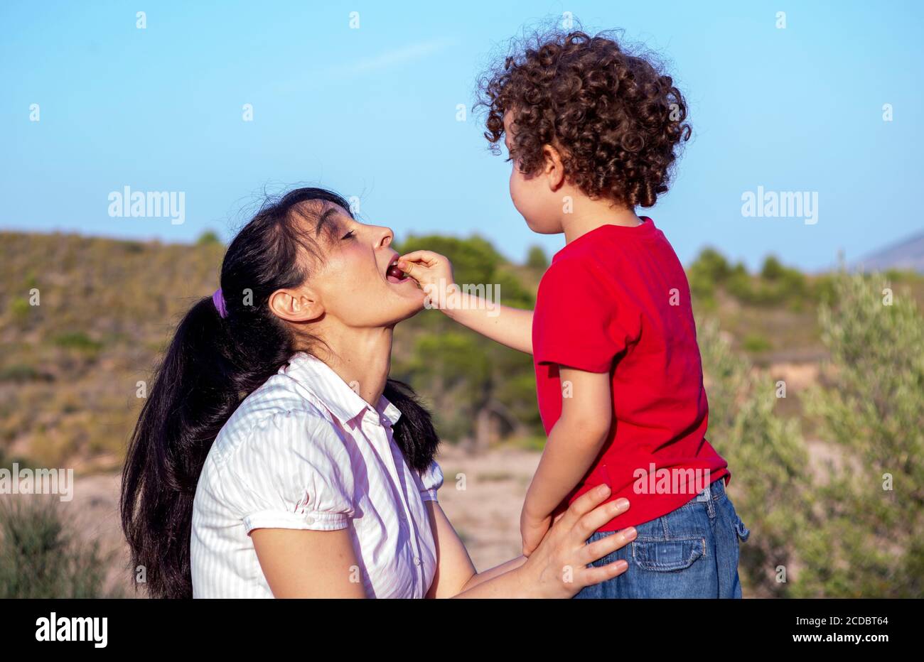 complicità e gioco tra madre e figlio che nutrono una ciliegia direttamente nella bocca della madre, godendo di una giornata di picnic in famiglia in montagna. Foto Stock