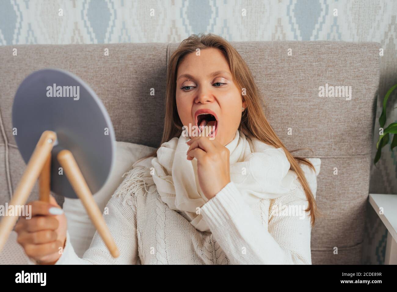 Bella femmina bionda controllare la gola con bastone di legno e. specchio sdraiato a letto e malato Foto Stock