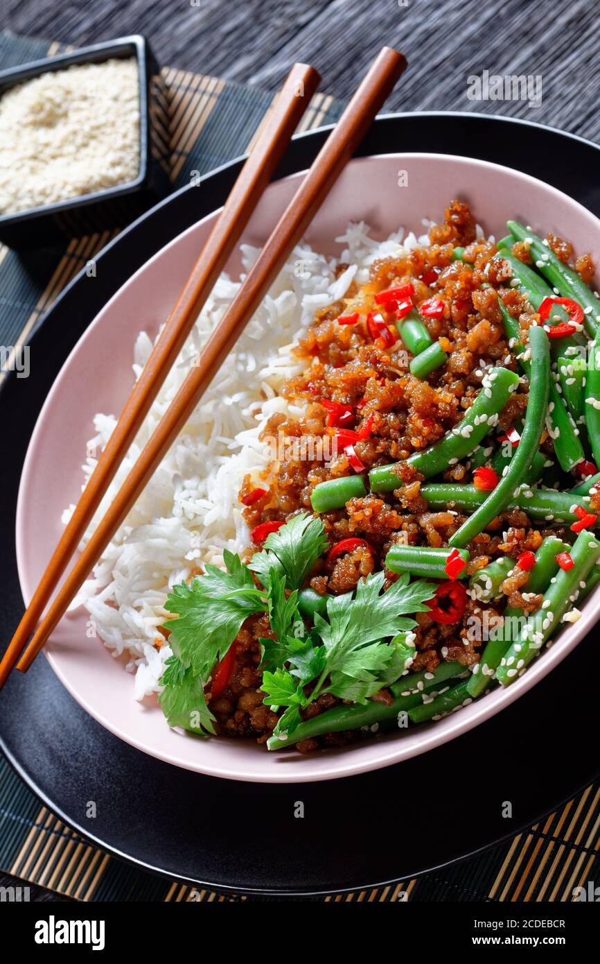 Primo piano su una ciotola rosa con zuppino di maiale tritato piccante asiatico kra pao con riso e fagioli verdi cosparsi di semi di sesamo, peperoncino rosso servito con chopstick Foto Stock