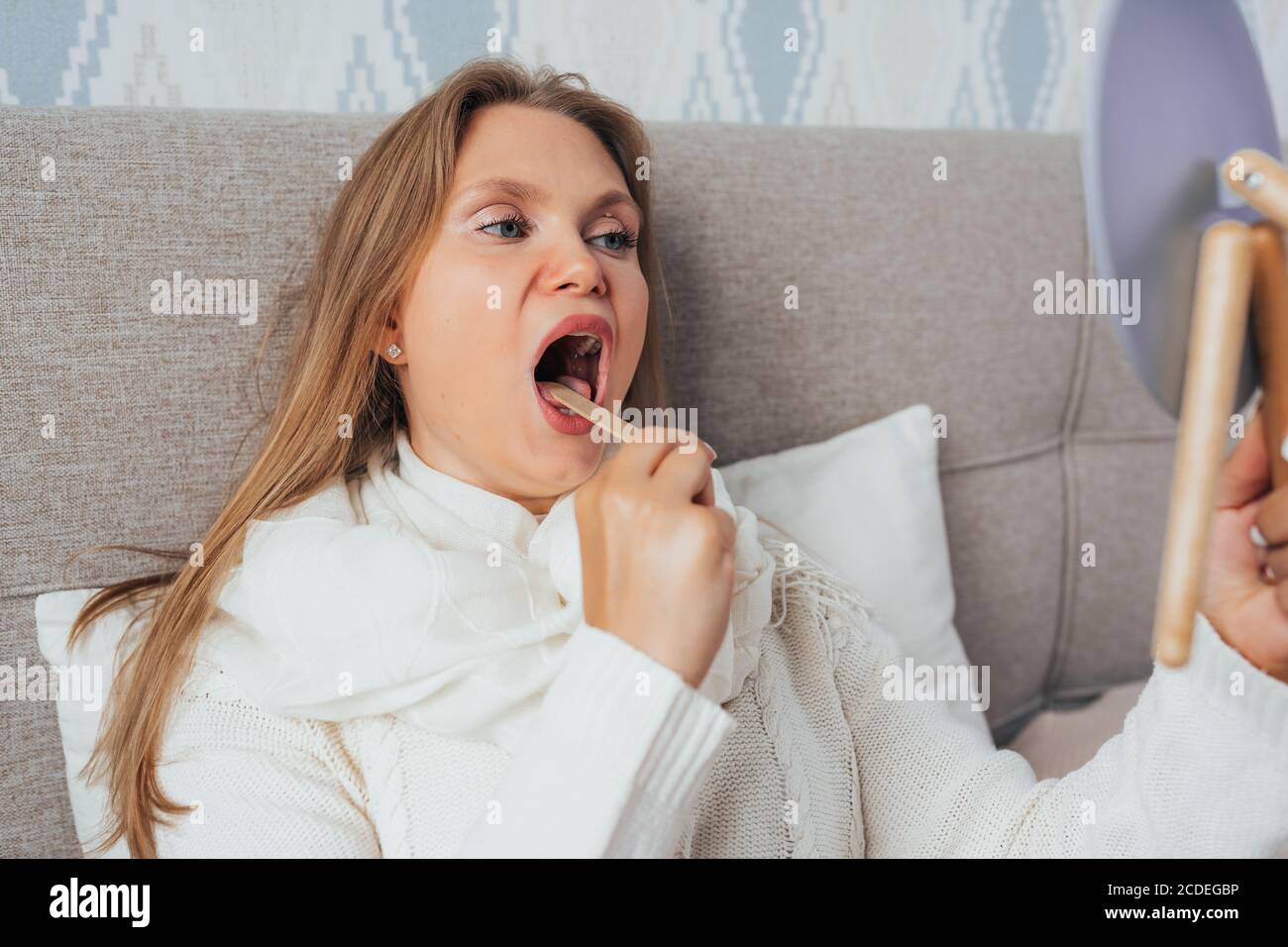 Bella donna bionda controllare la gola con bastone di legno e specchio sdraiato a letto e sentirsi malato. Foto Stock