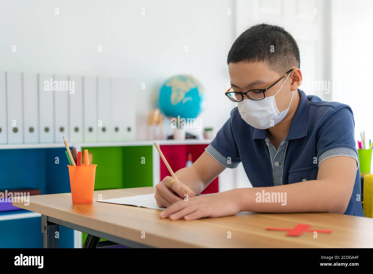 Studente asiatico di scuola elementare che indossa una maschera igienica per prevenire lo scoppio di Covid 19 in classe mentre torna a scuola riapre la sua scuola, New Foto Stock