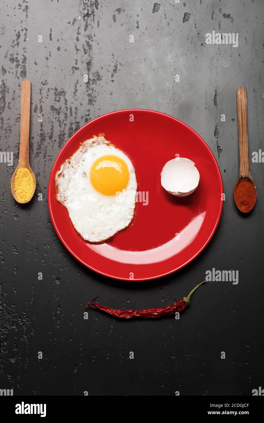 Colazione e concetto di arte alimentare. Omelette, gusci e riflessi fanno un'immagine sorridente del volto. Cibo posto su piatto rosso su fondo nero. Condire con uova fritte vicino a cucchiai con spezie e peperoncino rosso Foto Stock