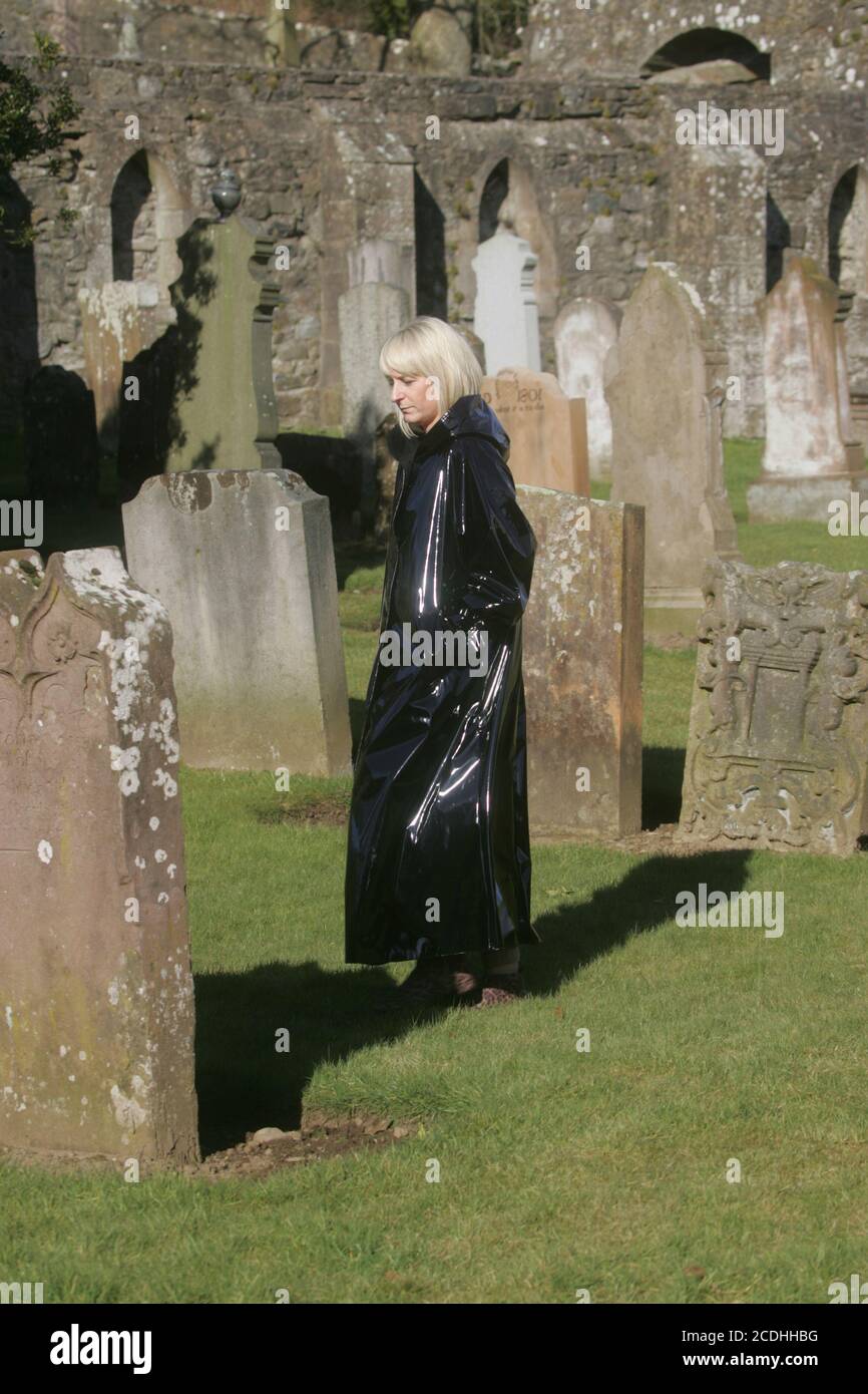 Kirkoswald, Ayrshire, Scozia, Regno Unito. Donna bianca caucasica in lungo lucido cappotto nero con cappuccio cammina intorno al cimitero in modo enigmatico Foto Stock