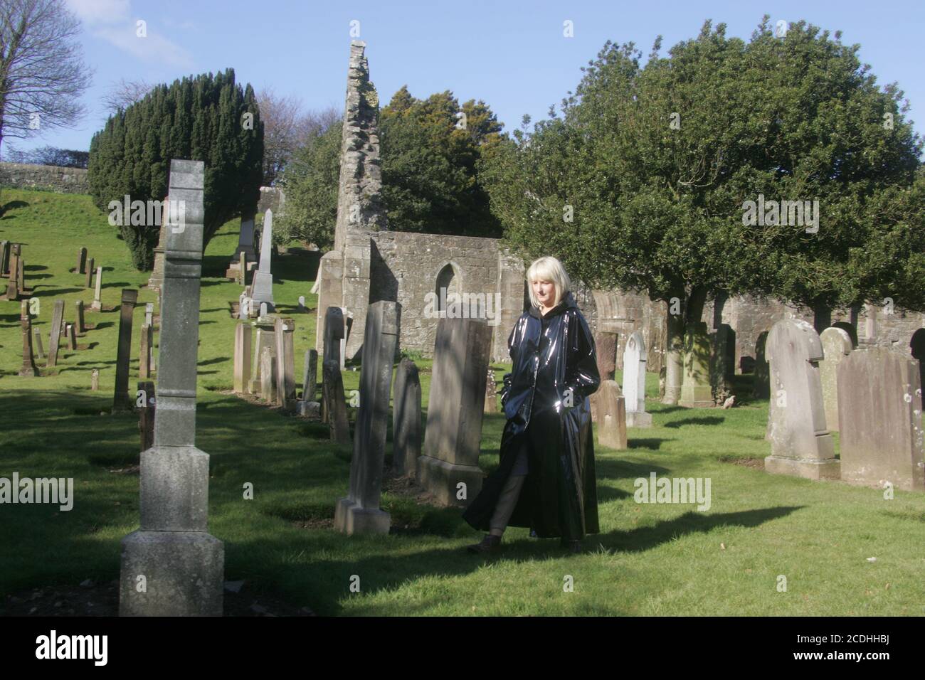 Kirkoswald, Ayrshire, Scozia, Regno Unito. Donna bianca caucasica in lungo lucido cappotto nero con cappuccio cammina intorno al cimitero in modo enigmatico Foto Stock