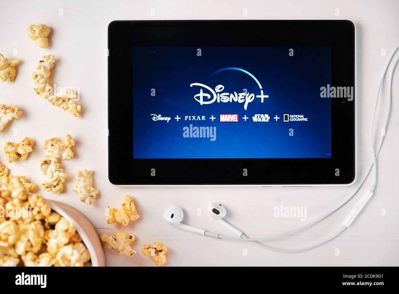 Il logo Disney Plus sullo schermo della tavoletta che si stette sul tavolo bianco e vi cosparse di popcorn. Auricolari Apple vicino al tablet che mostrano un Disney Foto Stock