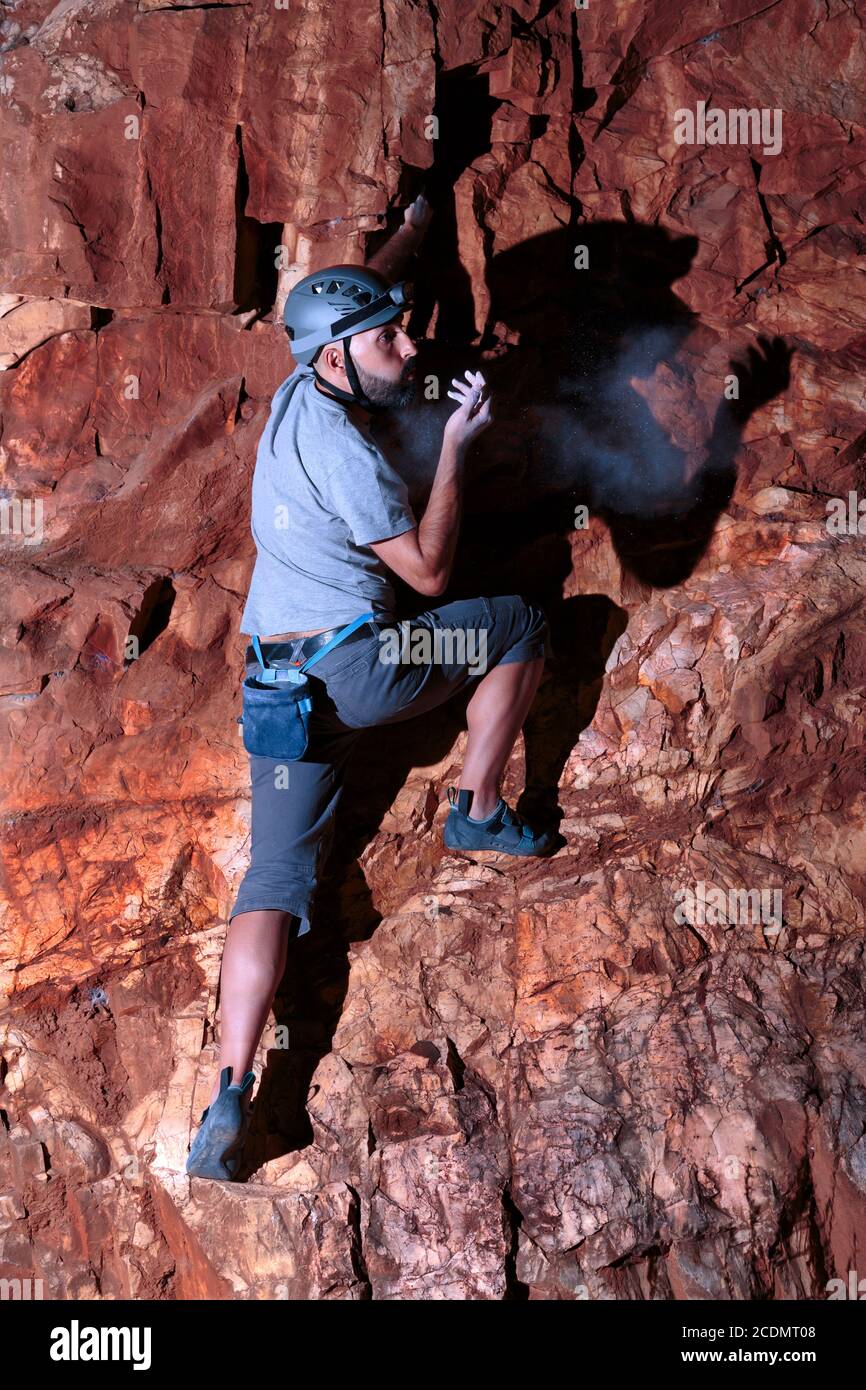 Abile scalatore ispanico maschio con la sua attrezzatura da arrampicata su, soffia il gesso in eccesso di polvere dalla sua mano destra mentre sale su un intenso muro di ferro rosso. Foto Stock