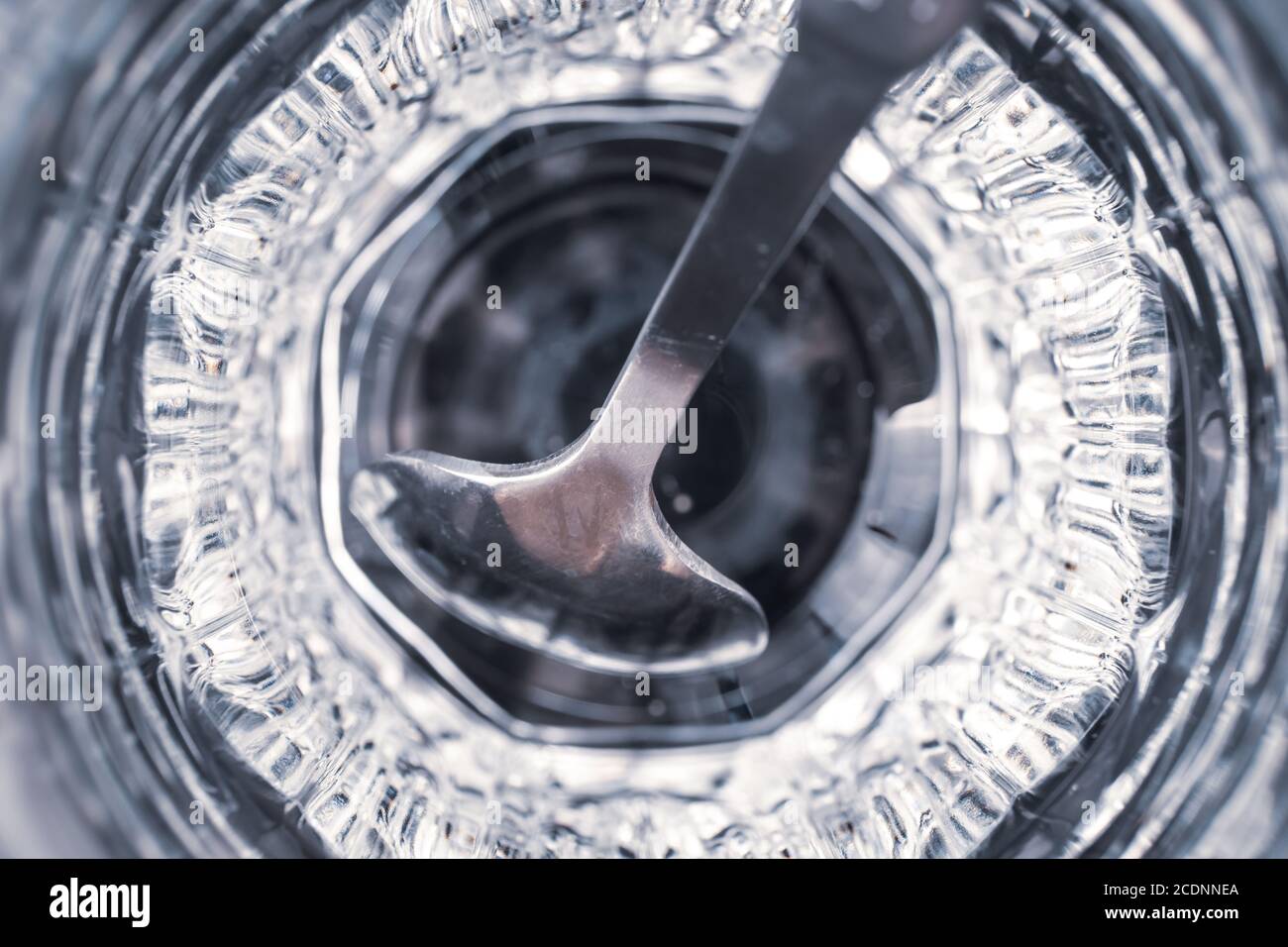 Cucchiaino da tè in metallo in una tazza di vetro sfaccettata con riflessi, vista dall'alto, primo piano. Sfondo astratto grigio e blu, sfondo futuristico, trasparente Foto Stock